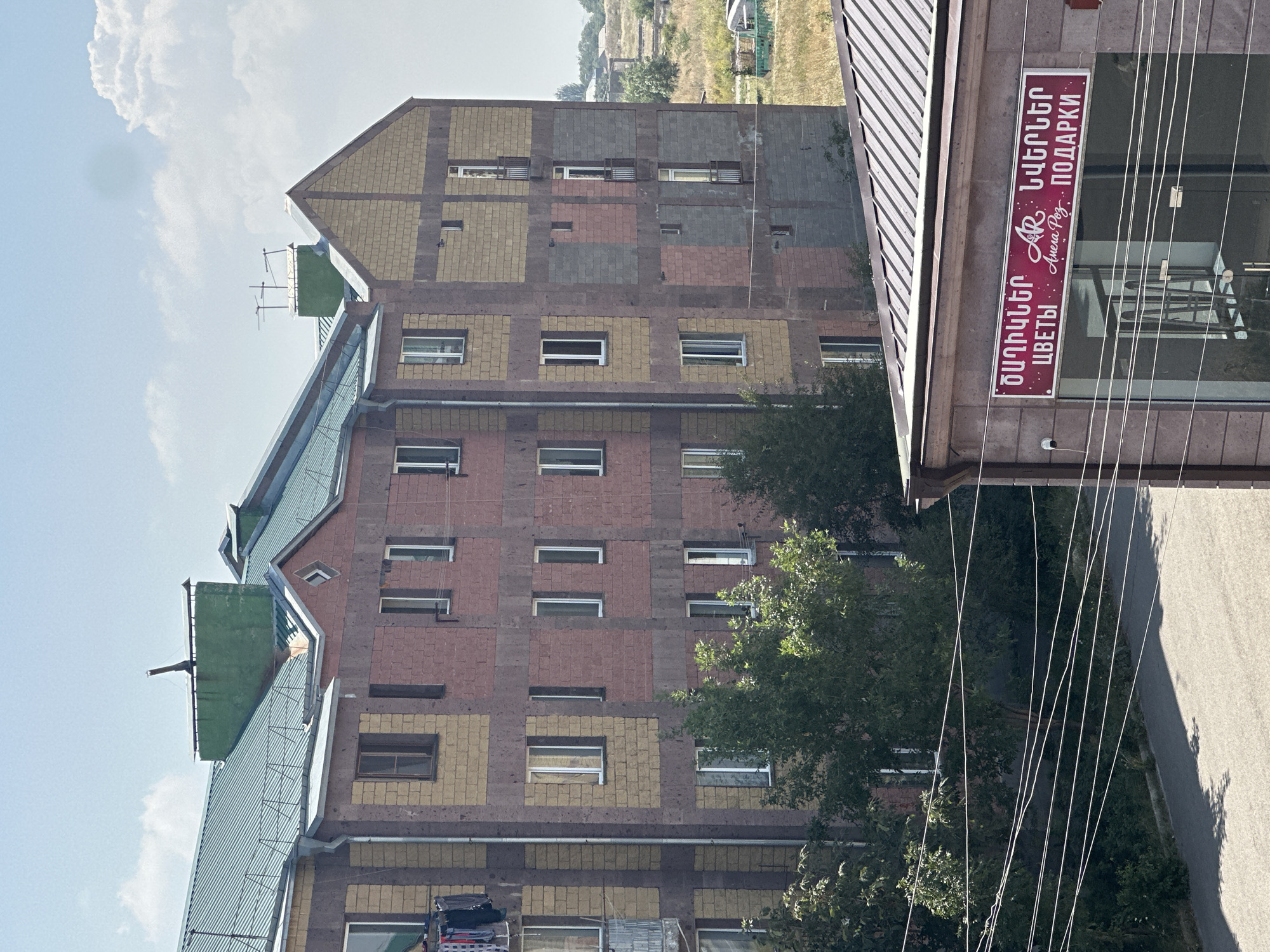 The image shows a view of a multi-story apartment building in Gyumri, Armenia. The building has a facade of pink and beige brick. There are several windows on each floor, some with clothes hanging from them. Trees are visible in front of the building, and a sign in Armenian and Russian indicates a shop below. The sky is blue with some clouds. The scene appears to be a residential area with a street in the foreground.