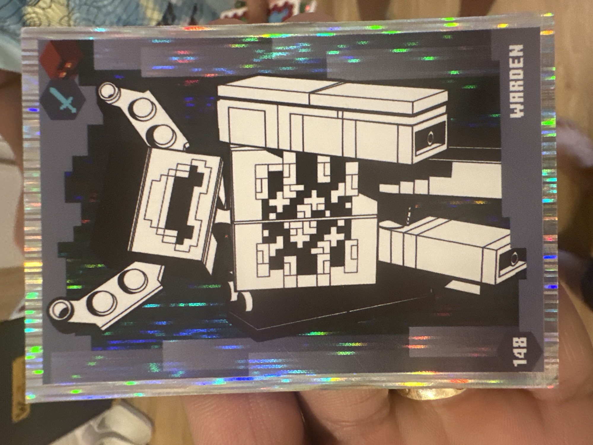 A person's hand is holding a holographic trading card featuring a stylized, pixelated robot character. The character, referred to as "Warden" by the text on the card, is predominantly black and white with a blocky design. The card itself has a holographic border with a rainbow effect. The background of the card depicts a dark, abstract city-like environment with vertical streaks of light. In the upper right corner, there is a small blue sword icon and a red block. The number "148" is visible in the lower left corner of the card. The hand holding the card has a gold ring on one finger. The environment outside the card is blurred, but appears to be an indoor setting with wooden surfaces. There is no information about the city of Mainburg, Germany in the image.