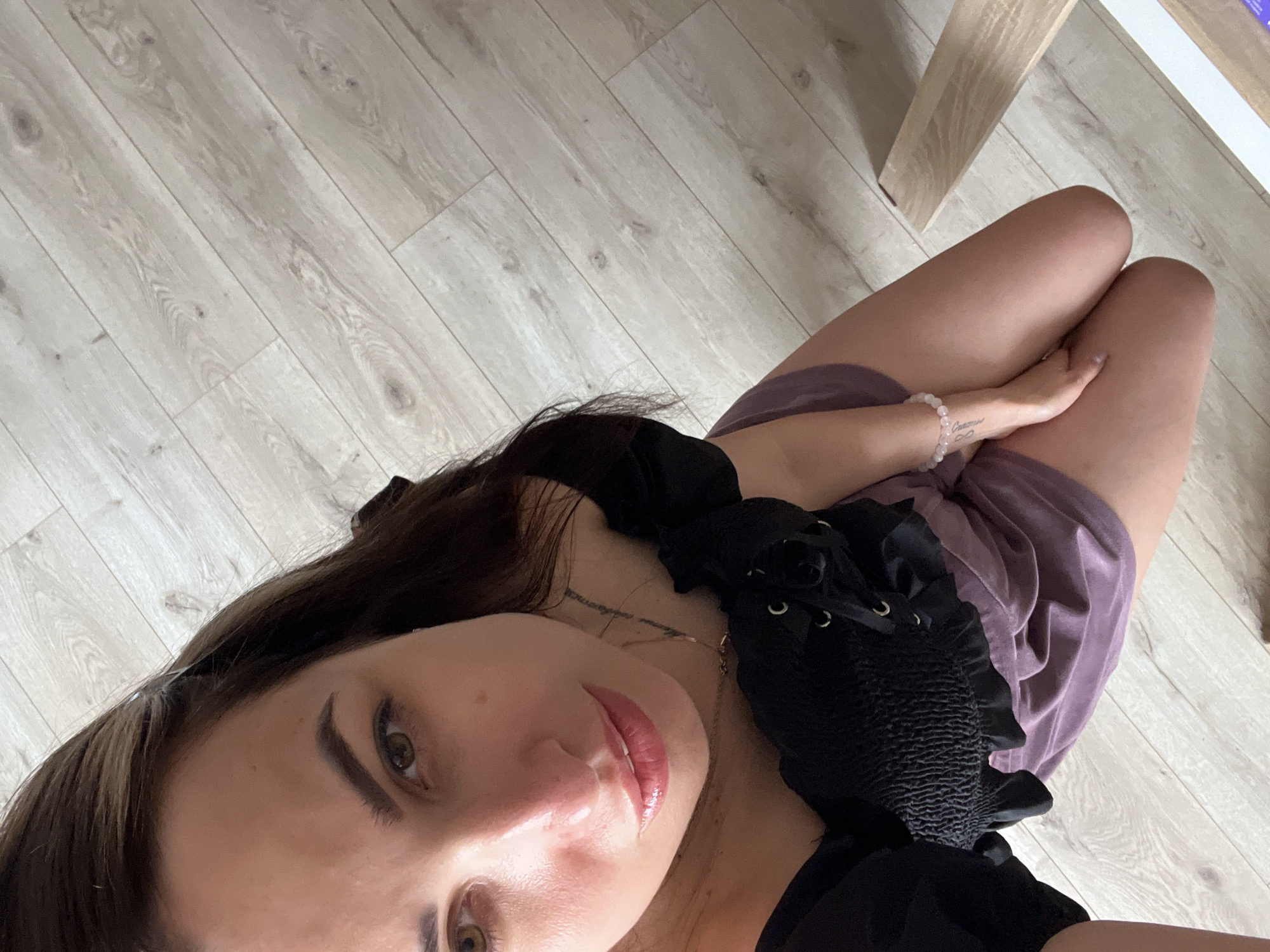 A woman kneels on a light-colored wooden floor. She is wearing a black off-the-shoulder top with a lace-up front and a dusty rose-colored skirt. Her hair is dark brown and falls past her shoulders. She is looking at the camera with a soft expression. A delicate necklace is visible around her neck, and a bracelet adorns her wrist. A tattoo is visible on her left forearm. The room appears to be lit by natural light.