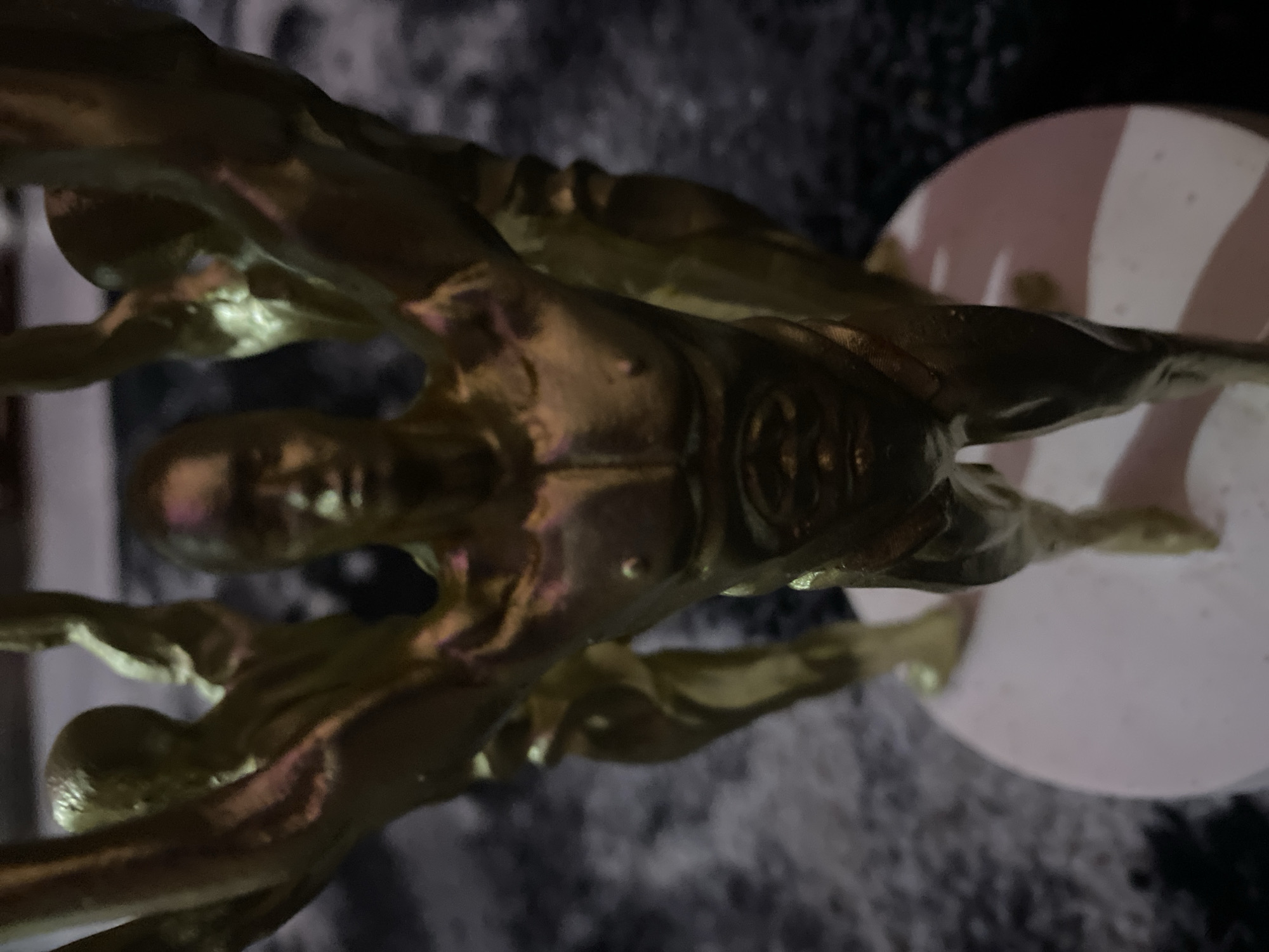The image displays a golden trophy with multiple sculpted male figures. The central figure is prominently featured, with arms raised. Behind and to the sides of the central figure are other figures, also appearing to be in a similar pose. The trophy is mounted on a circular, light-colored base. The background is dark and appears to be a textured surface. The lighting suggests an indoor setting, possibly with some ambient colored light reflecting off the trophy. There are no discernible details indicating the specific location in Kason, Nigeria.