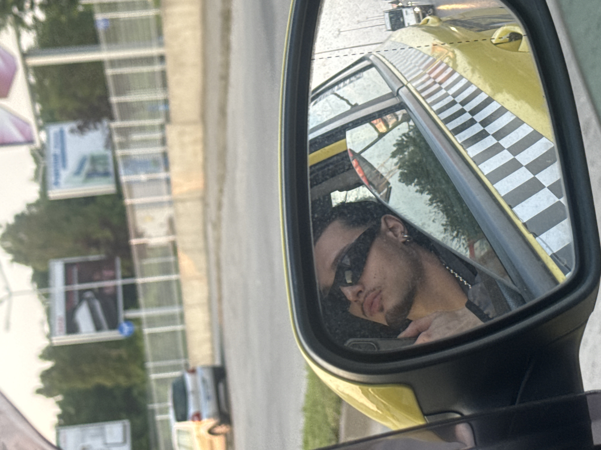 The image presents a detailed reflection within a yellow vehicle's side mirror, revealing a male occupant. The individual is wearing dark rectangular sunglasses, has a goatee, an earring in the visible ear, and a silver chain around his neck. A smartphone is partially visible in his hand near his face. The vehicle's exterior, visible in the mirror and extending to the left, is yellow and features a distinctive black and white checkered pattern along its side, identifying it as a taxi, consistent with those found in Sofia, Bulgaria.
Outside the mirror, the scene depicts a street or road environment under bright daylight. A light-colored hatchback car is observed on the road beyond the taxi. The background features a low concrete barrier, a metal fence, and a dense line of green trees. Above the trees, multiple billboards are visible; one on the right displays an advertisement featuring a white object against a dark background, with a blue circular sign positioned below it. Further down the road, in the far distance, a white vehicle resembling a bus or truck is also visible.