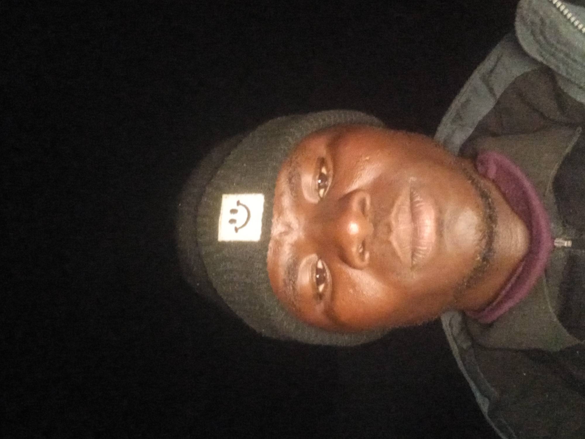 A close-up, front-facing, waist-up, selfie-style photograph features a dark-skinned man wearing a dark beanie with a white smiley face patch. He is wearing a dark jacket over a purple-collared shirt. The background is completely black, suggesting a dark environment, possibly at night. The lighting is focused on the man's face, creating harsh shadows. The image quality is low, with visible grain and motion blur. The man's expression is neutral. There are no discernible environmental features or context that would place the scene in Katon Rikkos, Nigeria or any specific city.