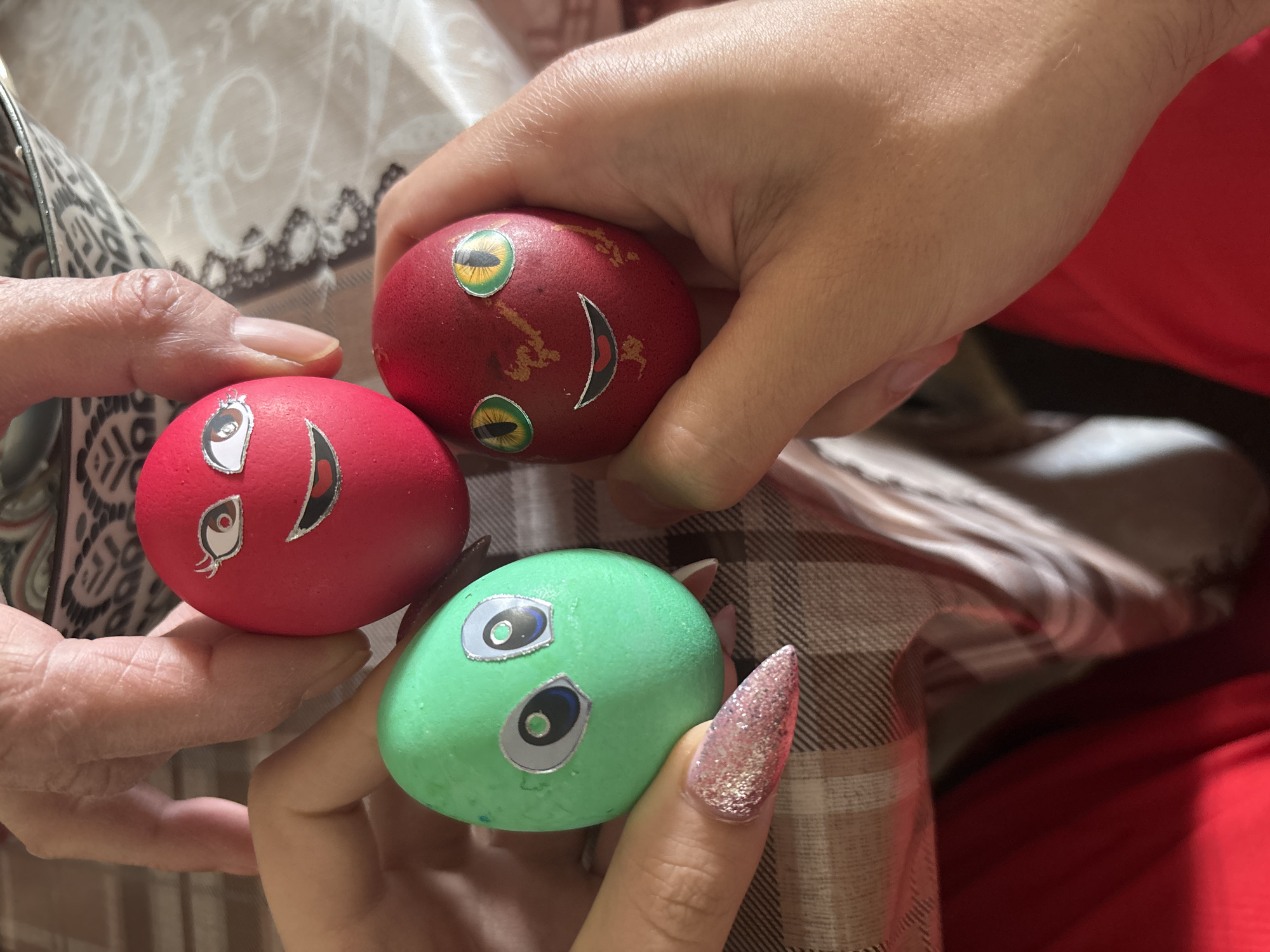 A close-up shot shows three hands holding three painted eggs, likely for Easter. One hand, on the left, holds a red egg. Another hand holds the red egg. A hand with a long, glittery fingernail holds a green egg. The red egg has a smiling face sticker. The green egg has alien eye stickers. The background includes a patterned cloth, and a red garment. The location is Goritsa, Bulgaria.