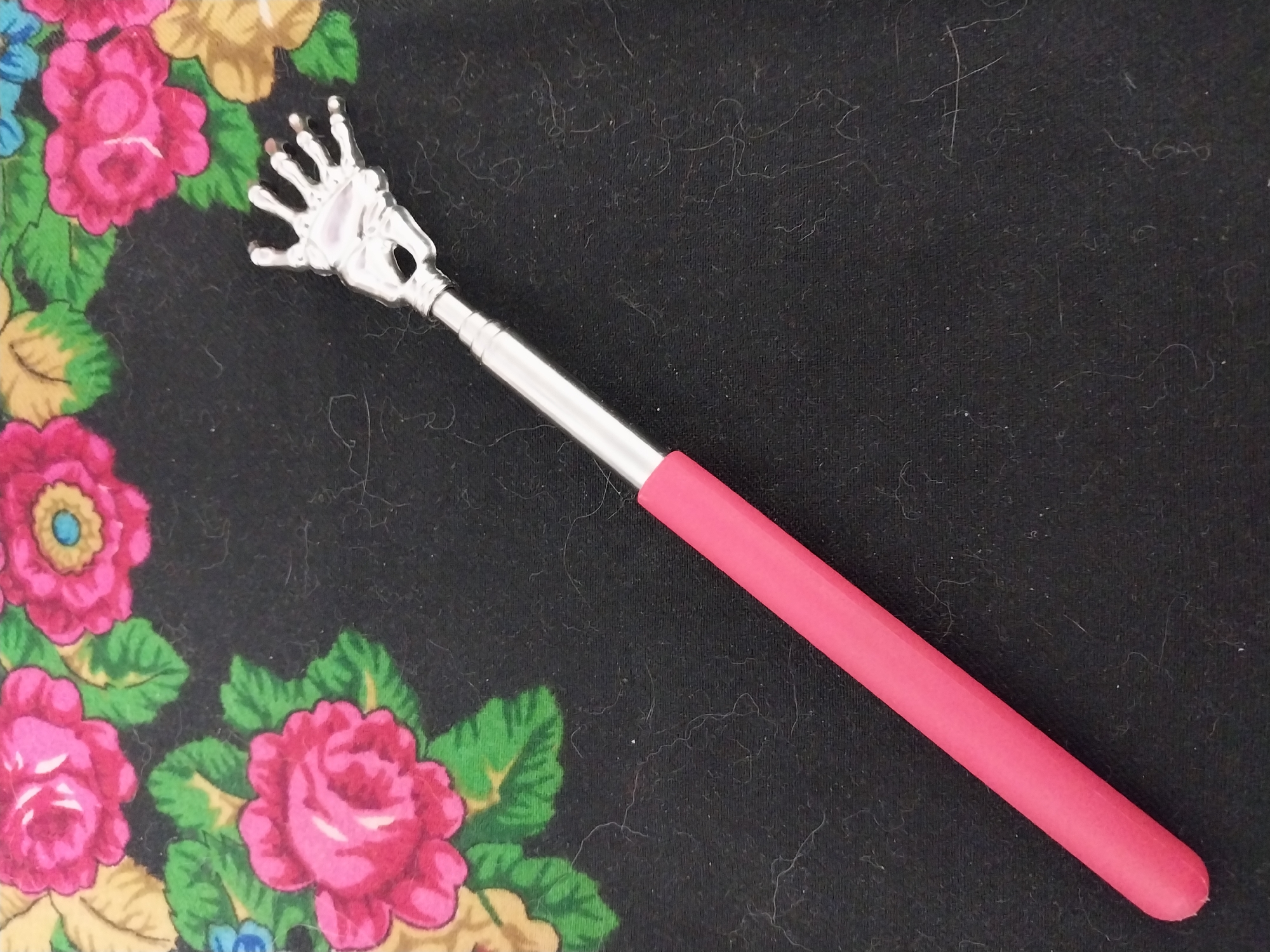 A metal back scratcher with a pink handle lies on a black fabric surface with a floral pattern. The back scratcher is shaped like a hand with outstretched fingers. The floral pattern features pink and yellow roses with green leaves. The scene is indoors.