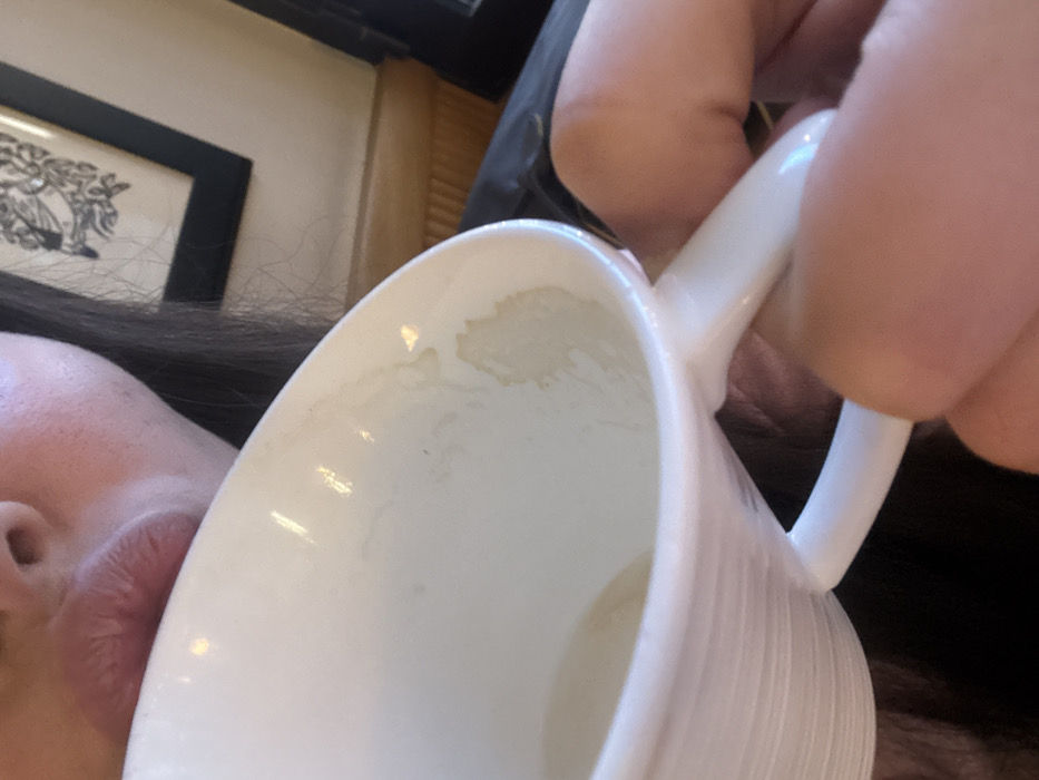 A white ceramic mug with a handle is held close to the face of a person with dark hair and fair skin. Residue lines the inside of the mug. The background includes a black-framed picture on a light-colored wall. A blurry hand holds the mug's handle.