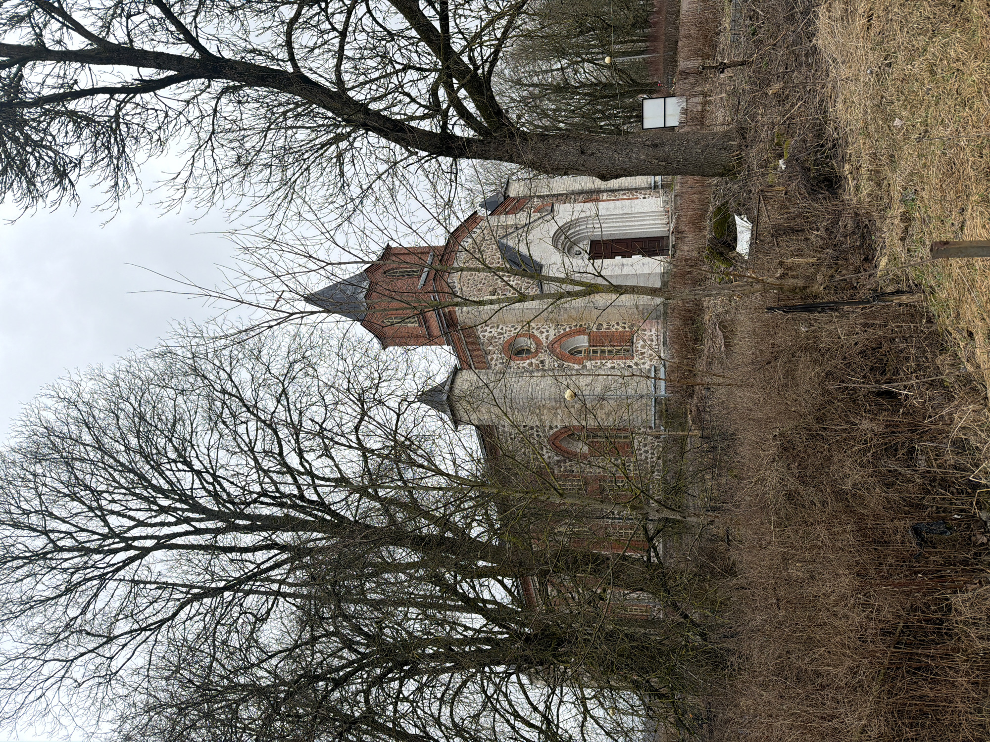 The image shows the Church of St. Nicholas in the village of Torosovo, Russia. It is a stone building with red brick accents, partially obscured by bare trees and overgrown foliage. The sky is overcast. No people are visible.