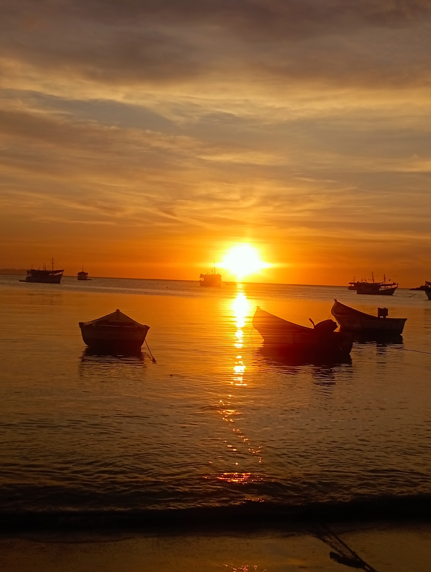 The image depicts a sunset over the ocean in Juan Griego, Venezuela. In the foreground, the beach is visible with small waves lapping at the shore. Several small boats are anchored in the water, their silhouettes reflecting the intense orange and yellow light of the setting sun. In the distance, larger ships are silhouetted against the horizon. The sky is filled with orange and yellow clouds, creating a warm and luminous atmosphere.