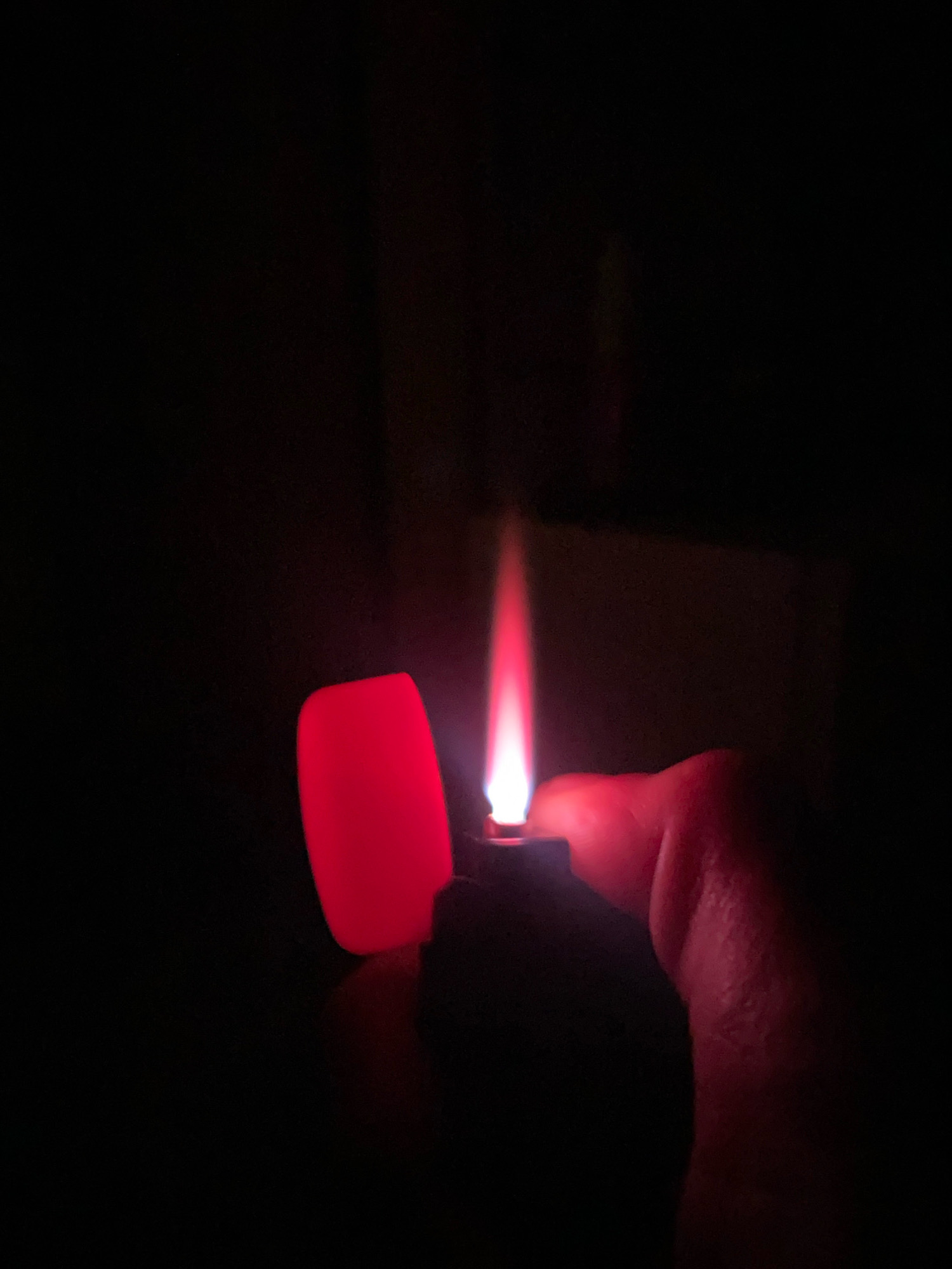 A close-up, medium shot captures a hand holding a red lighter with a lit flame against a dark background. The lighter's flame emits a bright, ethereal glow that illuminates the surrounding area with shades of red and orange. The hand, appearing to be that of an adult, is partially visible, with its skin reflecting the warm light of the flame. The lighter itself is predominantly red, with its cap open to reveal the mechanism. The overall ambiance is one of focused illumination in an otherwise dark environment. The location of Zarechny, Russia, is not discernible from the visual information provided in the image.