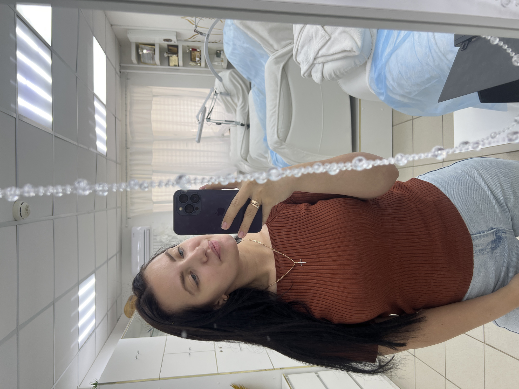 The image shows a young woman with long, dark hair taking a selfie in a mirror. She is wearing a rust-colored, ribbed short-sleeved shirt and light-wash jeans. A delicate cross necklace is visible around her neck. She is also wearing a ring on her right ring finger. The reflection in the mirror reveals a room that appears to be a beauty salon or a medical clinic. Several white examination beds, some covered with blue sheets, are visible. There are also shelves with awards and equipment in the background, and fluorescent lighting fixtures on the ceiling. The tiled floor suggests an indoor commercial space. The specific location in Chaikovsky, Russia is not identifiable from the visual elements alone, but the environment suggests a professional setting.