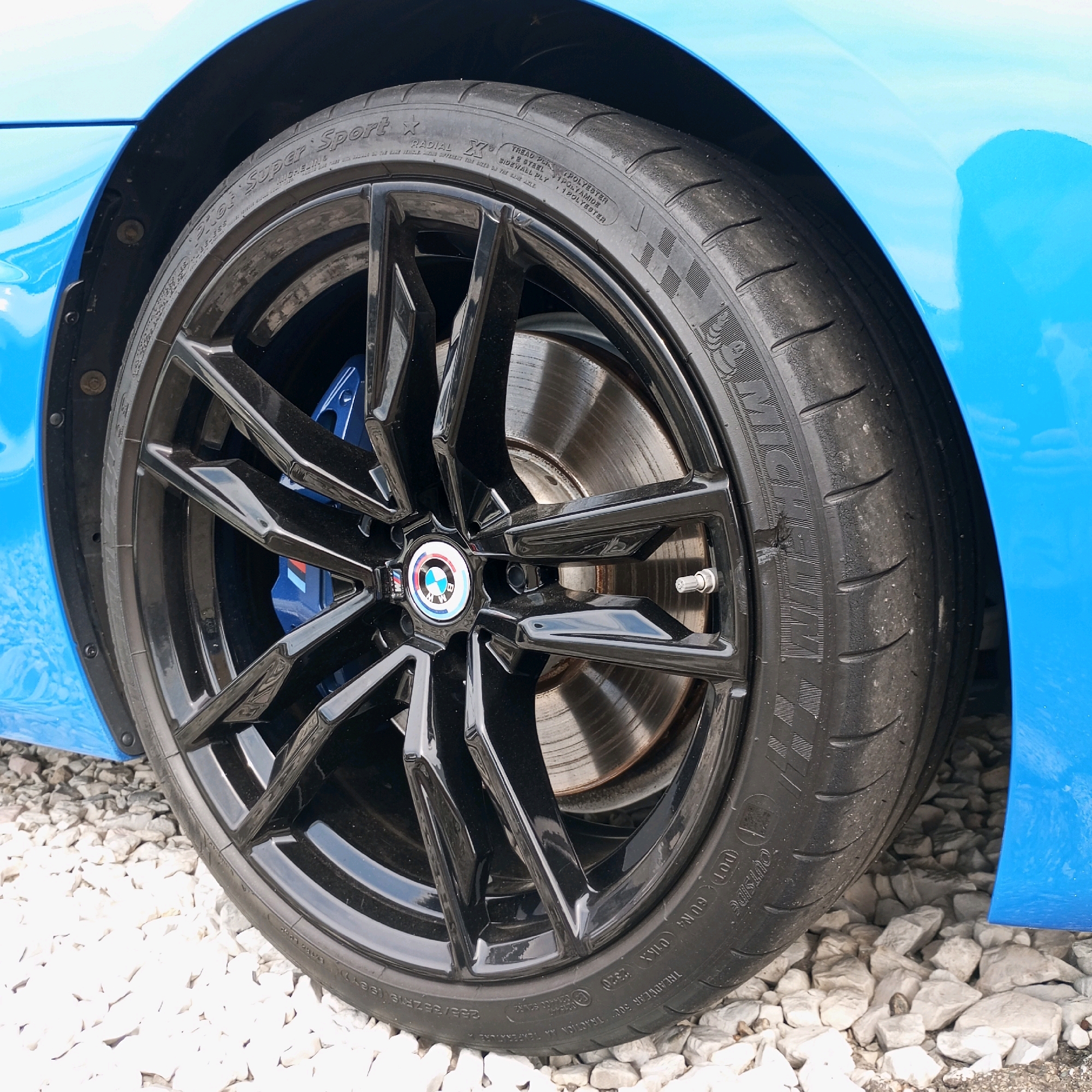 The image shows a close-up of a black alloy wheel on a bright blue car. The wheel has a complex, multi-spoke design and is fitted with a Michelin Pilot Super Sport tire. The tire has visible tread wear and some minor damage. The car's brake caliper, painted bright blue, is visible through the spokes of the wheel. The car is parked on a surface covered with white gravel. The background shows a portion of the blue car's fender and the edge of a black plastic fender liner. The scene is outdoors, likely in daylight, as indicated by the lighting and shadows. There are no people visible in the image. The context of Sochi, Russia is not evident from the visual information in the image.