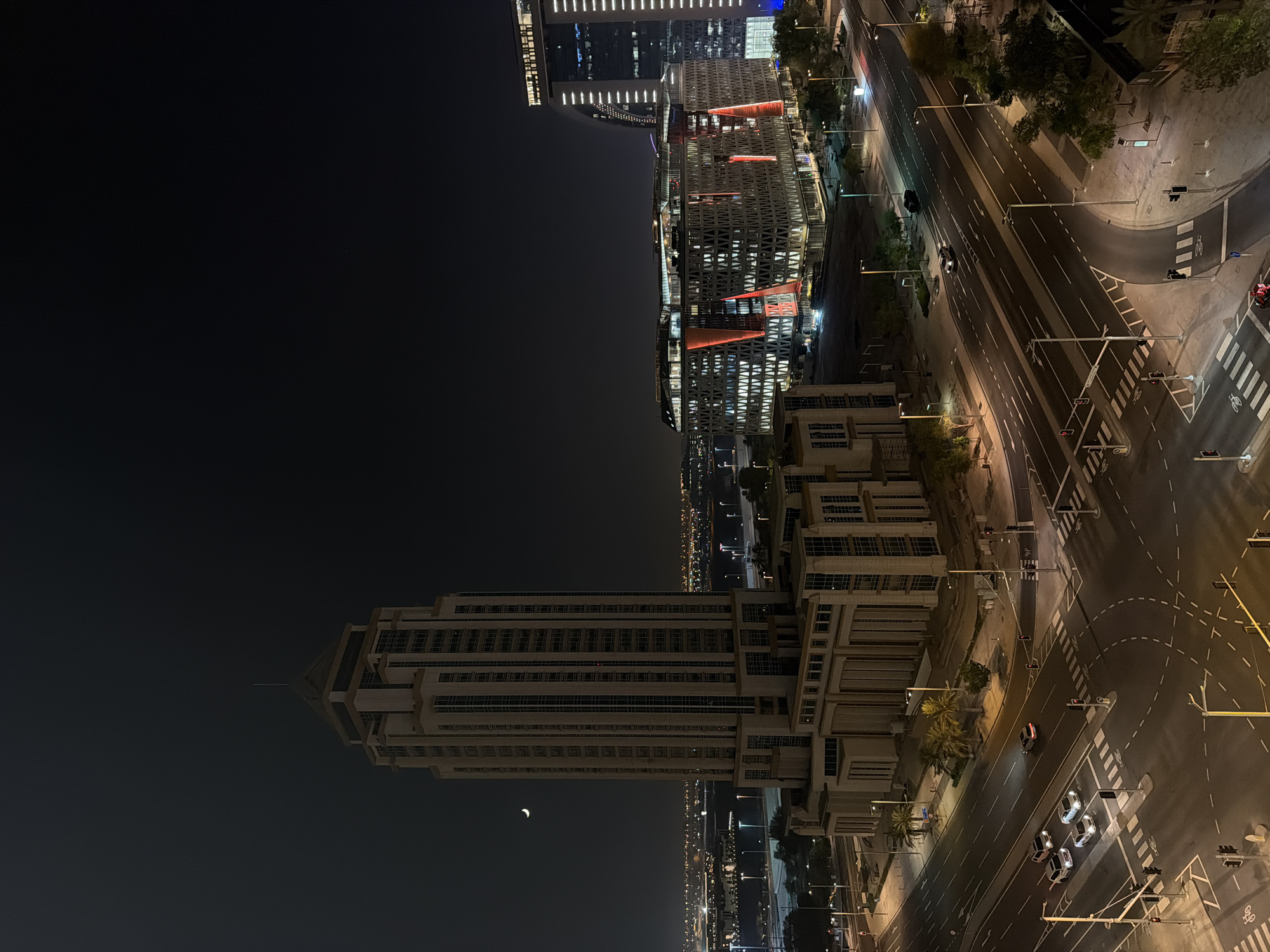 The image depicts a nighttime aerial view of a city intersection with several buildings and a crescent moon in the sky. The most prominent structure on the left is a tall, multi-story building with a stepped top and many windows. To the right of this building is a modern, block-like structure with a geometric facade, illuminated with red and white lights. Further to the right, another tall building is visible, with a curved design and illuminated windows. The intersection below is a multi-lane road with traffic lights, pedestrian crossings, and several cars driving or stopped. Streetlights illuminate the roads and sidewalks, and some palm trees are visible. In the distance, the cityscape extends with numerous lights from other buildings and infrastructure. The sky is dark, with the crescent moon clearly visible in the upper left quadrant.