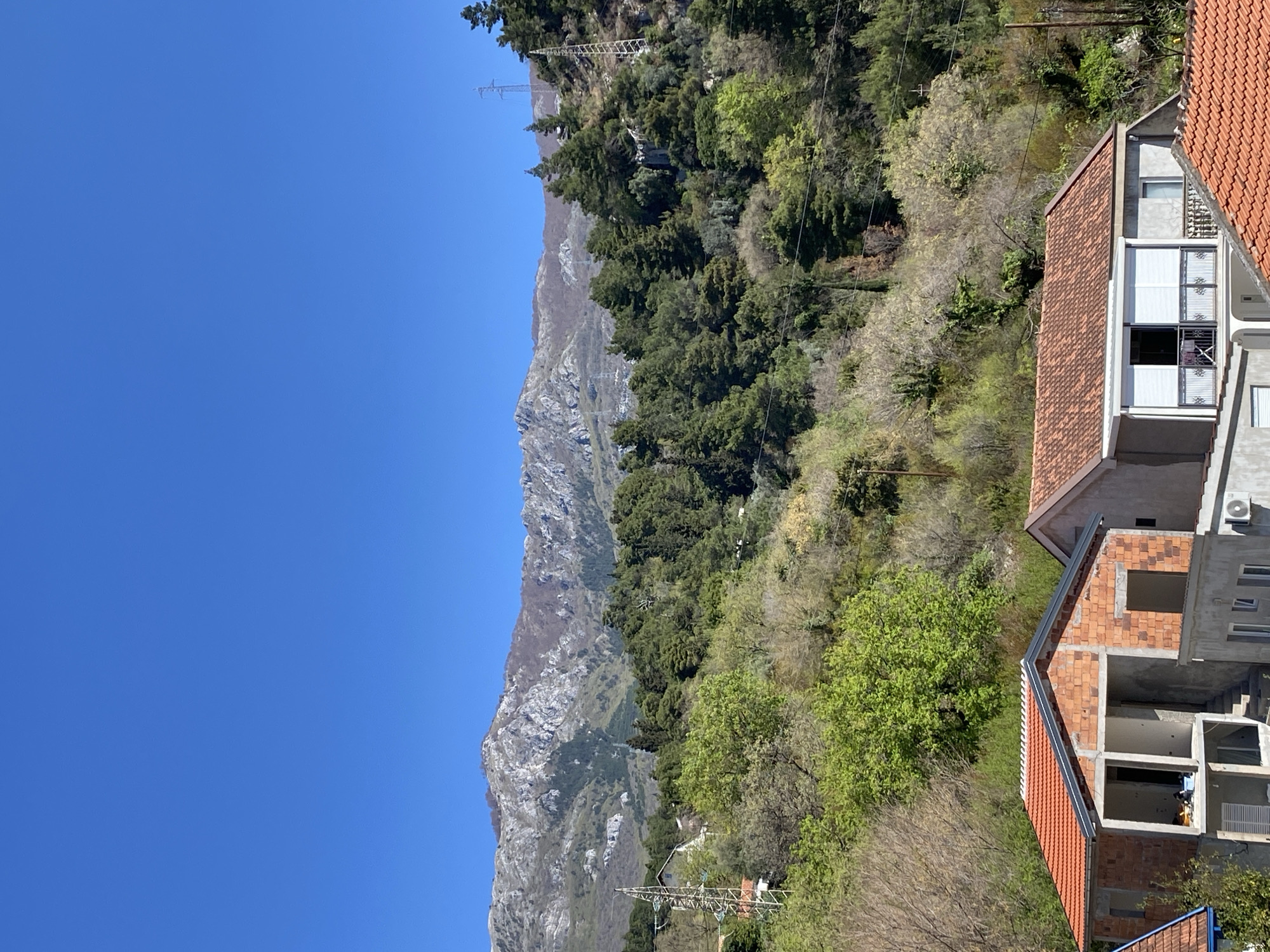 The image captures a scene in Budva, Montenegro, featuring a partial view of the cityscape against a mountainous backdrop. The sky is a clear blue. On the right side of the image, two buildings with red-tiled roofs are visible. One is partially constructed, exposing its brick framework, while the other appears more complete with finished windows and an external air conditioning unit. Lush green vegetation, including trees and shrubs, covers the hillside between the buildings and the mountain. Power lines run along the hillside. In the background, a rugged mountain range dominates the horizon.