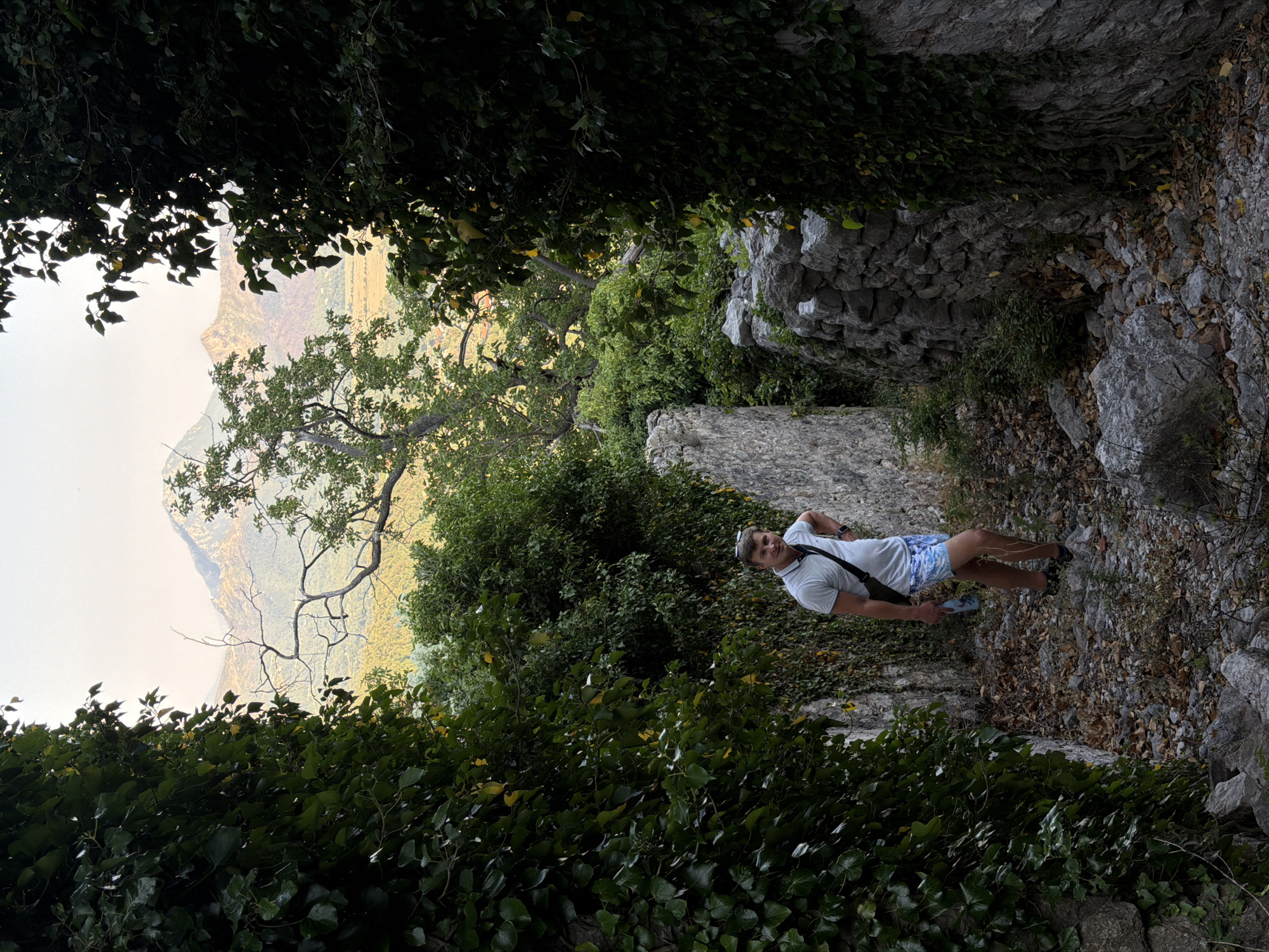 A man stands in a stone pathway overgrown with ivy and foliage. The man is wearing a light-colored polo shirt, patterned shorts, and dark shoes. He has a dark bag slung across his body. He is holding a water bottle in his left hand. The pathway is rocky and strewn with dry leaves. In the background, mountains are visible under a clear sky. The location is Androvići, Montenegro.