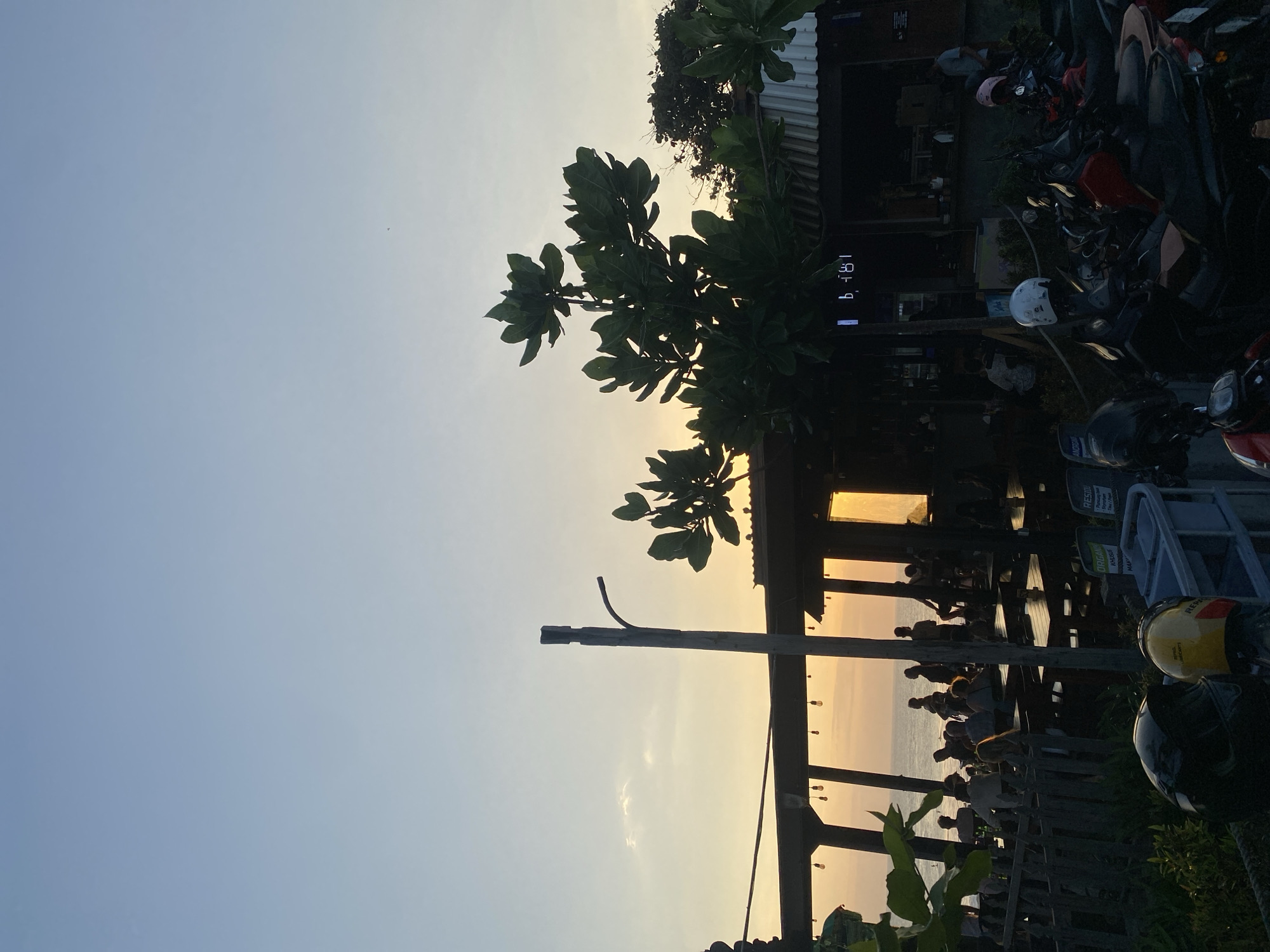 The image depicts a beachfront cafe or restaurant in North Kuta, Indonesia, during sunset. The sky is a gradient of pale blue to warm orange, with a hint of clouds near the horizon. The foreground is dominated by a collection of parked motorcycles, many of them scooters, with helmets visible on some. Behind the motorcycles, a wooden fence separates the viewer from the dining area.
The dining area itself is covered by a rustic, open-air structure supported by wooden beams. Tables and chairs are set up, and several groups of people are seated, appearing to be dining or socializing. The setting sun casts a warm glow through the open sides of the establishment and illuminates the sea in the background.
A prominent feature in the mid-ground is a large, leafy tree, its silhouette dark against the sunset sky. To the left of the tree, a tall, slender pole with a bent arm, likely a lamp post, stands out. Further into the building, illuminated signs and equipment suggest a functional establishment. The overall atmosphere is one of a relaxed, casual evening gathering by the coast.