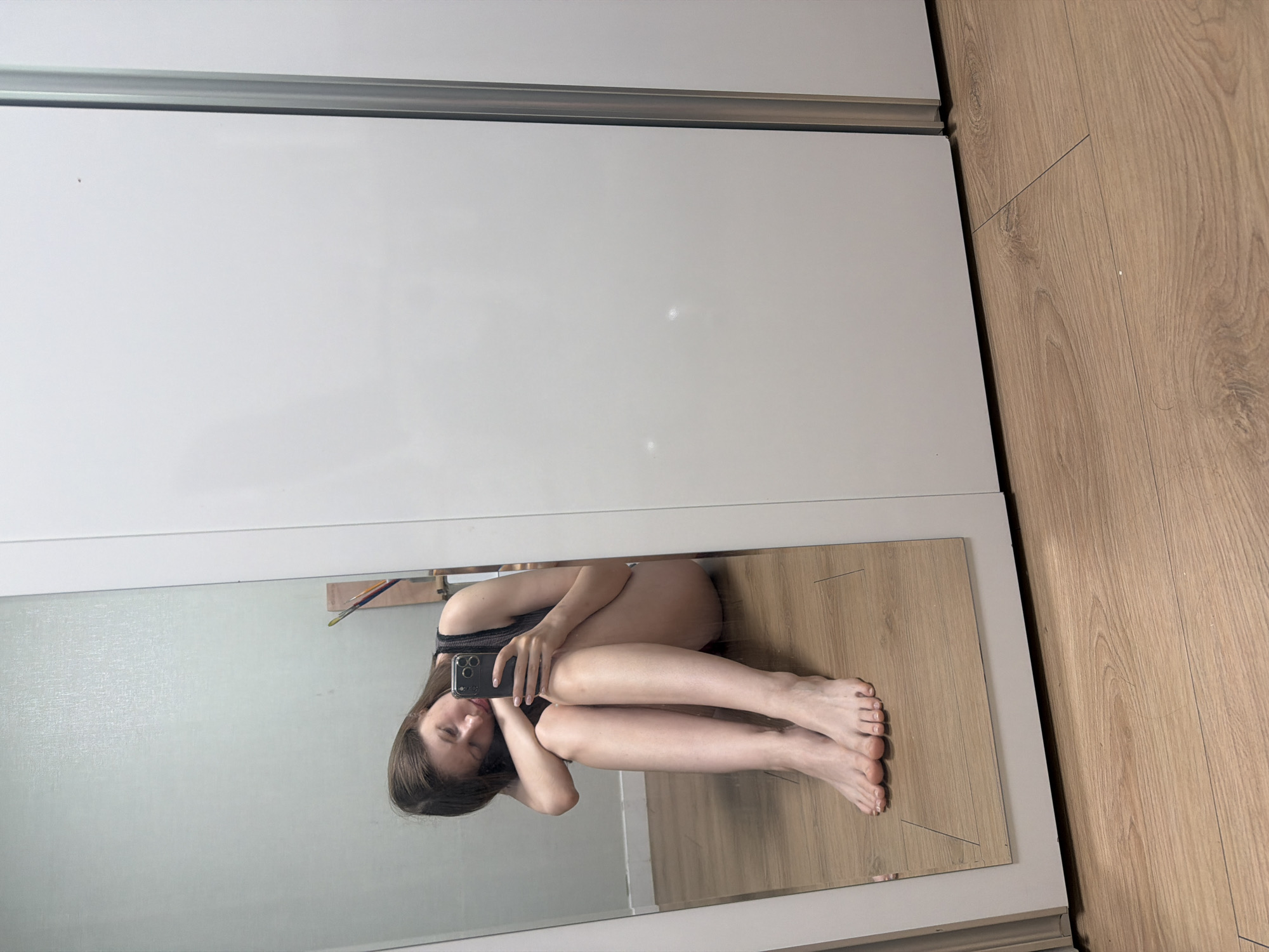 A young woman with long brown hair is sitting on a wooden floor, facing a mirror. She is holding a smartphone in her right hand and is taking a selfie. Her eyes are closed, and her head is resting on her left arm, which is bent at the elbow. Her legs are crossed, and her feet are visible in the mirror. She is wearing a dark-colored tank top and dark shorts. The background shows a wall and a closet with white doors. The scene appears to be indoors, in what could be a bedroom or a dressing room. There are no visible indications of the location being specifically in Dongducheon-si, South Korea.