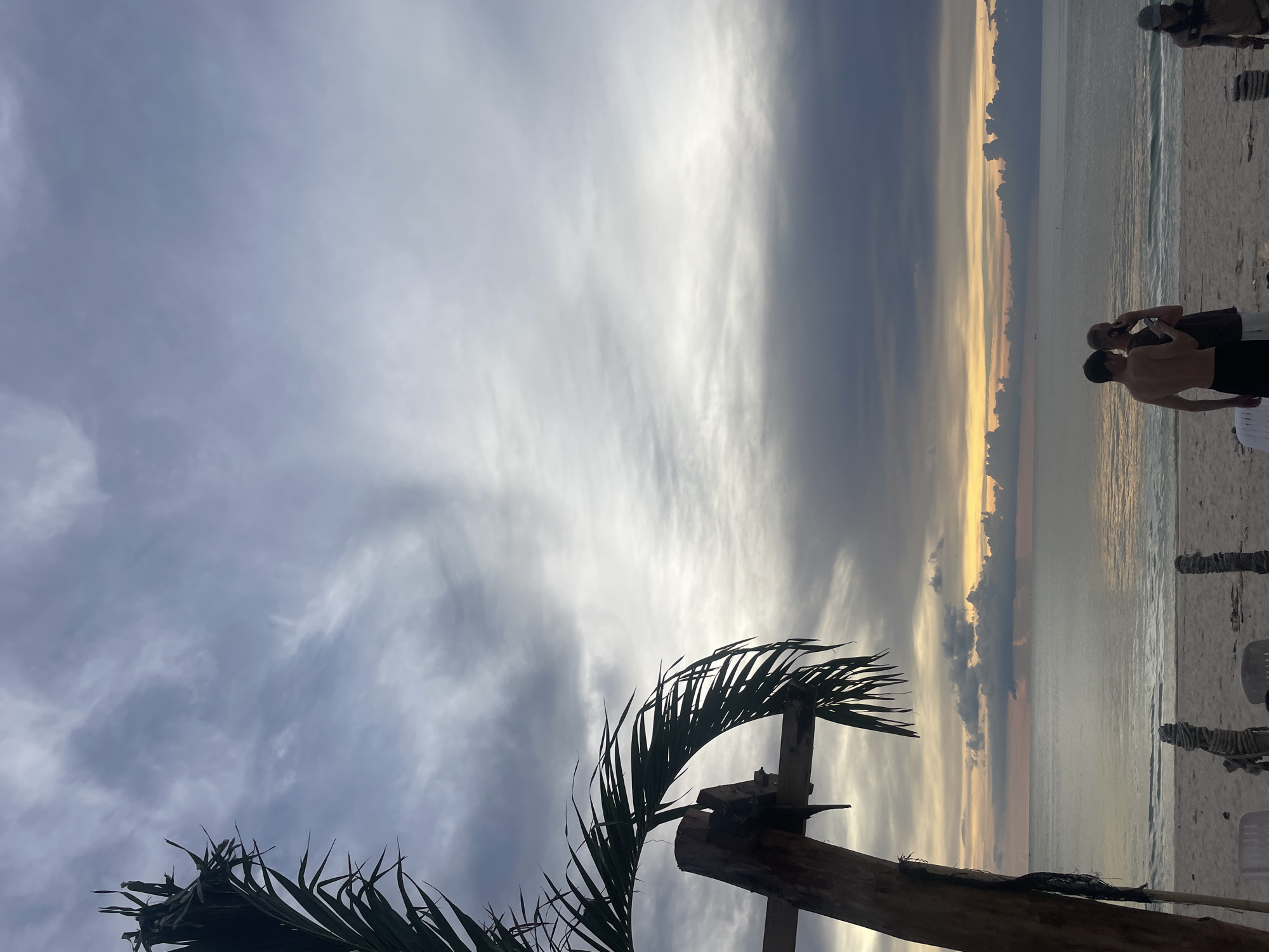 The image depicts a beach scene in Choeng Thale, Thailand. The sky is cloudy with visible sunlight peeking through the horizon. In the foreground, a portion of a palm tree and a wooden structure are visible. Two individuals are standing on the beach, one appearing to take a photo of the other, with additional individuals and beach chairs visible in the periphery. The sea stretches out towards the horizon, reflecting the colors of the sky.