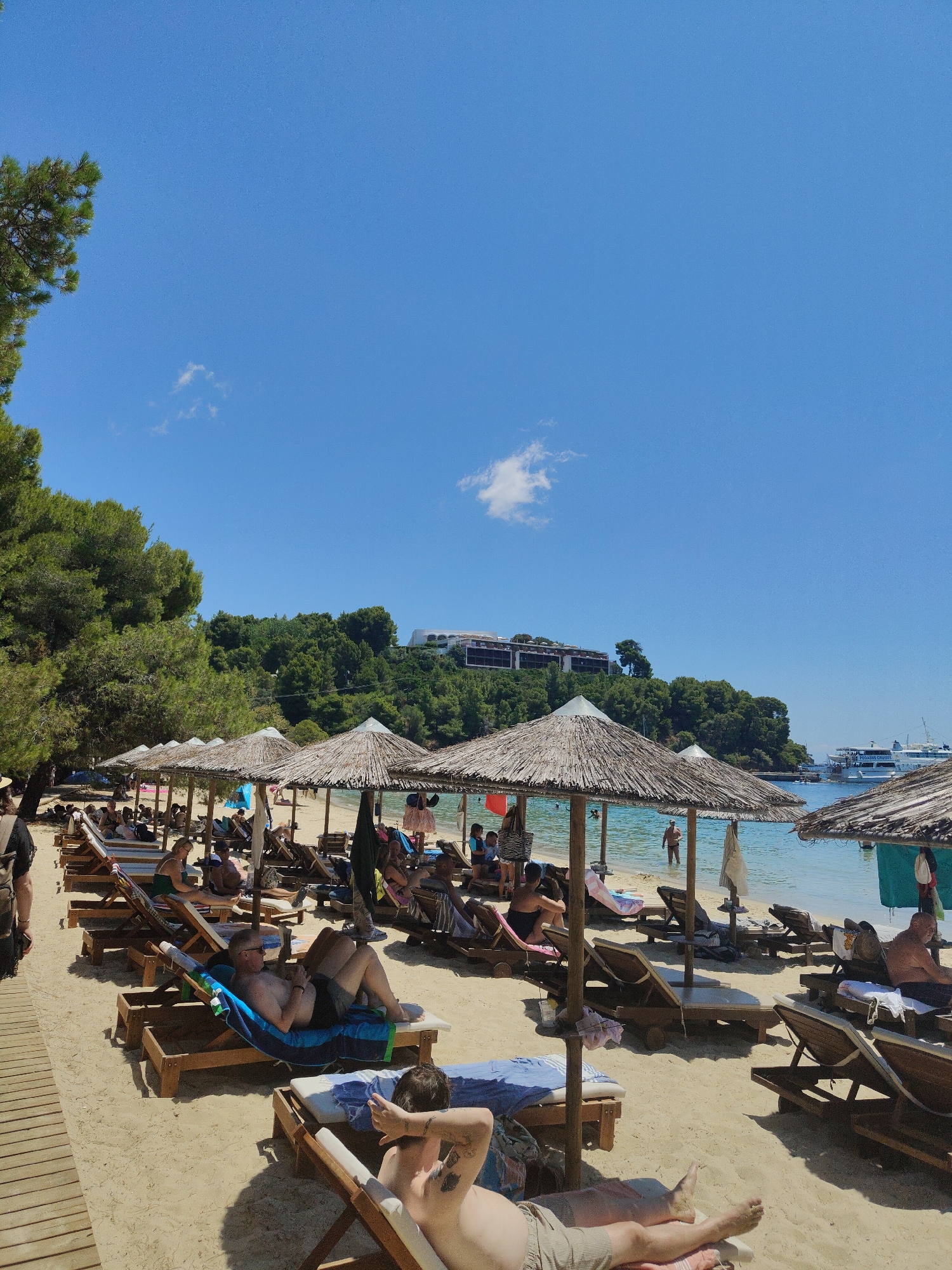 This is a medium-shot, eye-level, outdoor, daytime photograph of a crowded beach scene in Unknown, Greece. The foreground is dominated by rows of empty and occupied lounge chairs under thatched umbrellas on a sandy beach. In the middle ground, people are relaxing on the lounge chairs, some reading, some sunbathing, and some appearing to be looking at their phones. Further back, the clear blue water of the sea is visible, with several boats anchored offshore. Lush green trees and buildings are visible on a hillside in the background, under a bright blue sky with a few scattered clouds. The overall atmosphere is one of leisure and relaxation.
