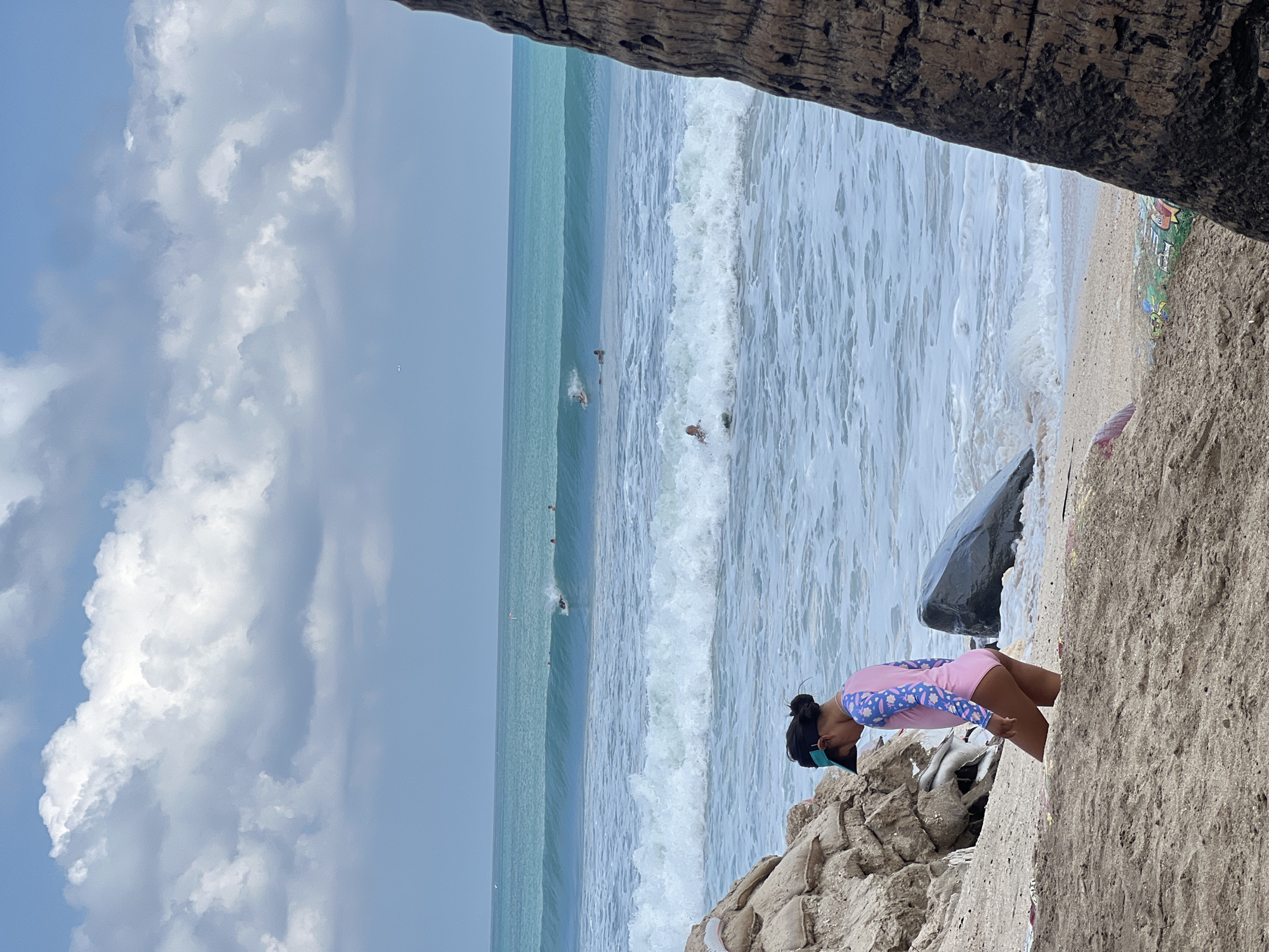 The image captures a beach scene in Kuta, Indonesia. A young girl wearing a pink swimsuit with blue sleeves is positioned on a sandy embankment, looking down at the rocks in front of her. She has her hair pulled back and is wearing goggles. Behind her, the turquoise ocean stretches out with waves breaking on the shore. Several people are visible in the water, some appearing to be swimming or surfing.
The sky is partly cloudy, with large, fluffy white clouds against a pale blue backdrop, suggesting it is daytime. The overall lighting indicates it is likely midday or early afternoon. The foreground is dominated by the rough texture of rocks and sand, framing the view of the ocean. The presence of a large dark rock to the left of the girl and the weathered wooden frame at the top of the image add to the natural and somewhat rugged coastal environment. There is some colorful graffiti visible on the sandy area further down the beach, adding a touch of artistic expression to the scene. The mood appears relaxed and summery, with the girl seemingly absorbed in her surroundings.