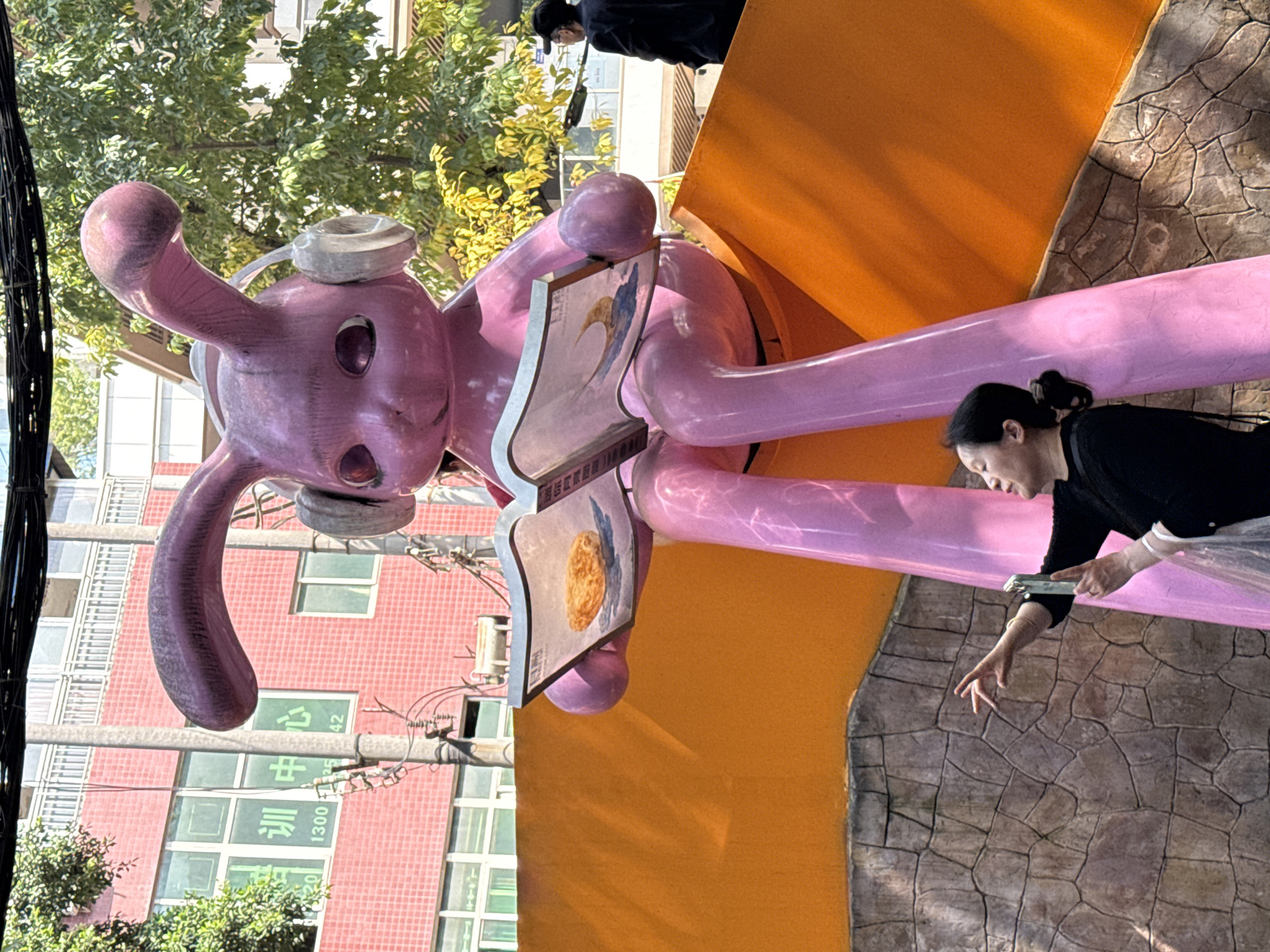 The image, rotated 90 degrees clockwise, presents an outdoor scene in Nan'an District, China, on a bright, sunny day. A large, glossy pink sculpture, resembling a stylized rabbit or long-eared creature, dominates the foreground. It is positioned horizontally on a bright orange structure, wearing silver headphones and holding two open "books" or screens. One screen clearly displays a golden, textured object, possibly a cookie or moon, against a blue and white background, with Chinese characters below.
In the mid-ground, a woman in a black top, with her hair tied back, is seen reaching out with her right hand, fingers splayed, towards the ground or the sculpture. She is positioned near one of the pink sculpture's extended limbs, above a grey, stone-textured surface. Further in the background, another person in dark attire is partially visible near the orange structure.
The backdrop features lush green trees and a multi-story red-brick building with numerous windows. Text visible on the building includes the Chinese characters "训练中心" (xùnliàn zhōngxīn, meaning "training center") and numbers such as "1300" and "42". Overhead, intricate black power lines and cables are strung across the sky. The strong daylight casts distinct shadows, highlighting the vibrant colors of the sculpture and the surrounding environment.