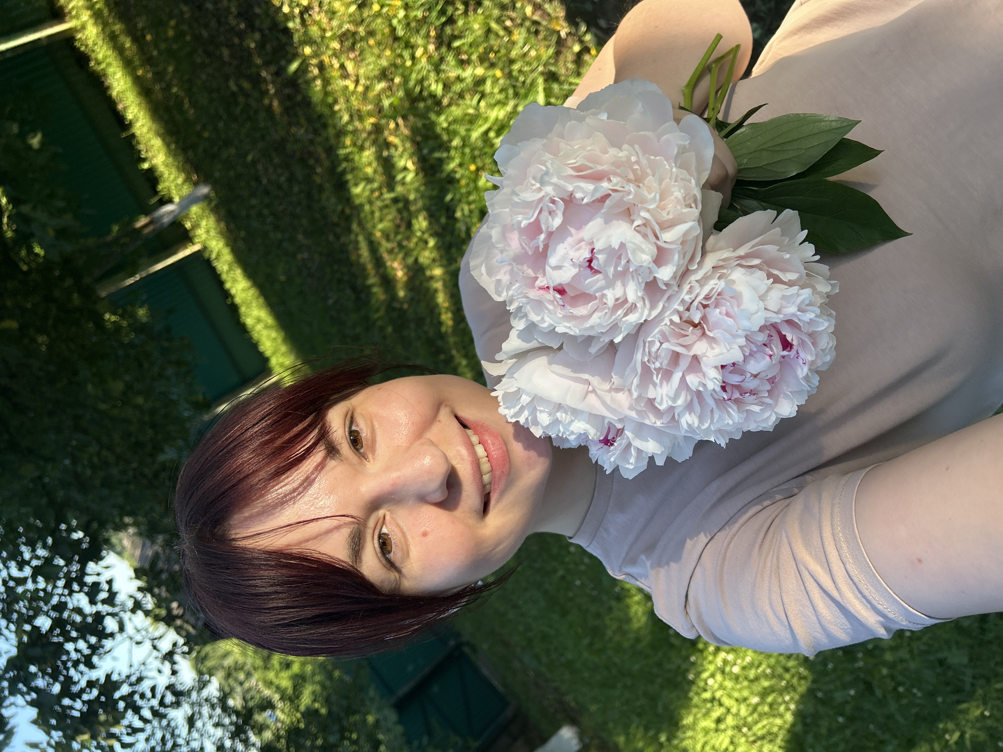 In the city of Foky, Russia, a young woman with auburn hair and bangs is captured in a selfie outdoors. She is smiling and holding a bouquet of three large, light pink peonies with darker pink centers. She is wearing a light tan t-shirt. The background shows a lush green lawn bathed in sunlight, with dappled shadows from trees. A dark green fence is visible in the background, along with the trunks of trees. The scene suggests a bright, sunny day, possibly in a garden or park.