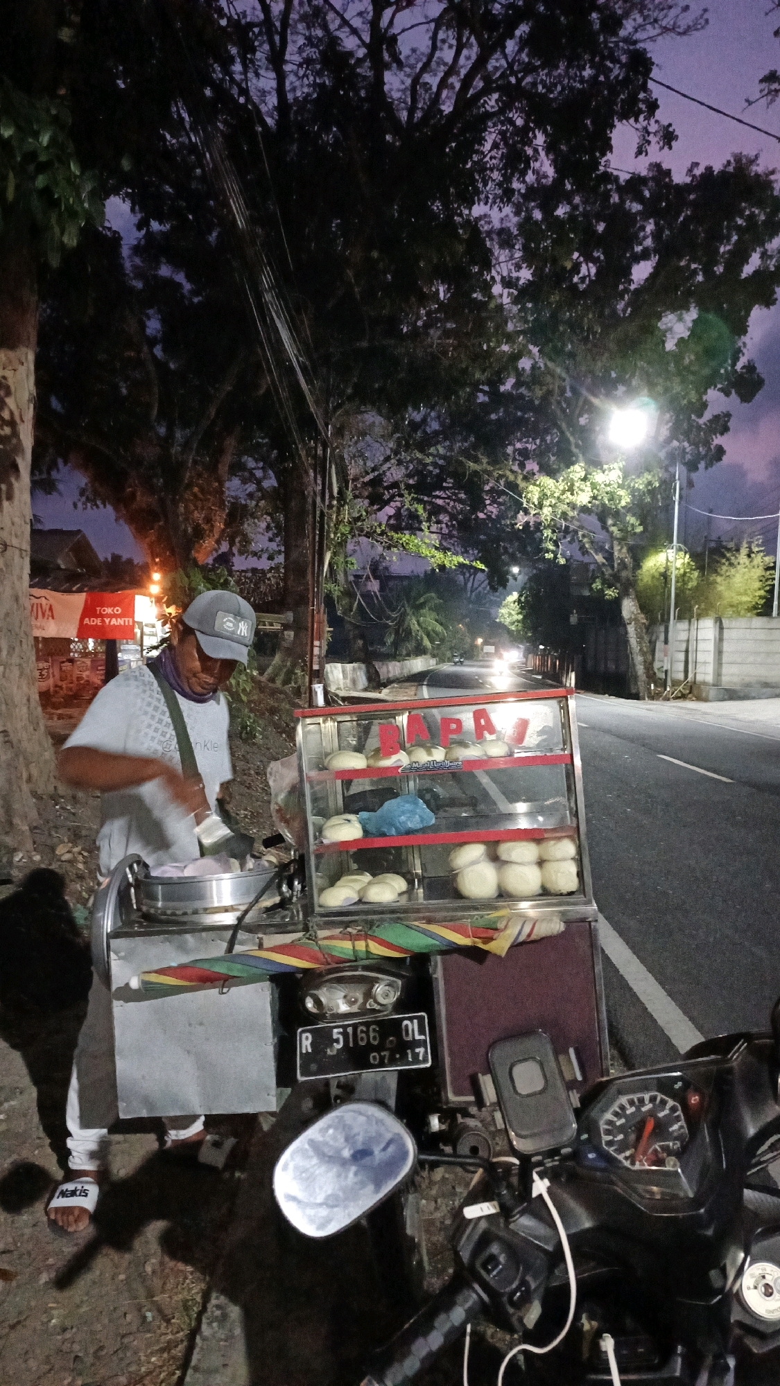 In the city of Sukaresik, Indonesia, a street vendor is preparing steamed buns at dusk. The vendor, wearing a gray t-shirt, gray baseball cap, and a bandana around their neck, is actively engaged in their work behind a steaming cart. The cart displays a variety of round, light-colored buns on multiple shelves within a glass enclosure. A blue plastic bag is also visible on one of the shelves.
The scene unfolds on the side of a paved road, with a motorcycle parked to the right of the vendor. The motorcycle's dashboard, including the speedometer, is visible, as is a license plate bearing the inscription "R 5166 OL 07-17." The surrounding environment is characterized by tall trees with dense foliage and a twilight sky, hinting at the transition from day to night. Streetlights illuminate parts of the scene, casting a warm glow. In the background, traffic can be seen on the road, indicated by illuminated headlights. A building with signage is partially visible to the left of the vendor. The ground beneath the vendor and the cart appears to be rough, possibly dirt or gravel.