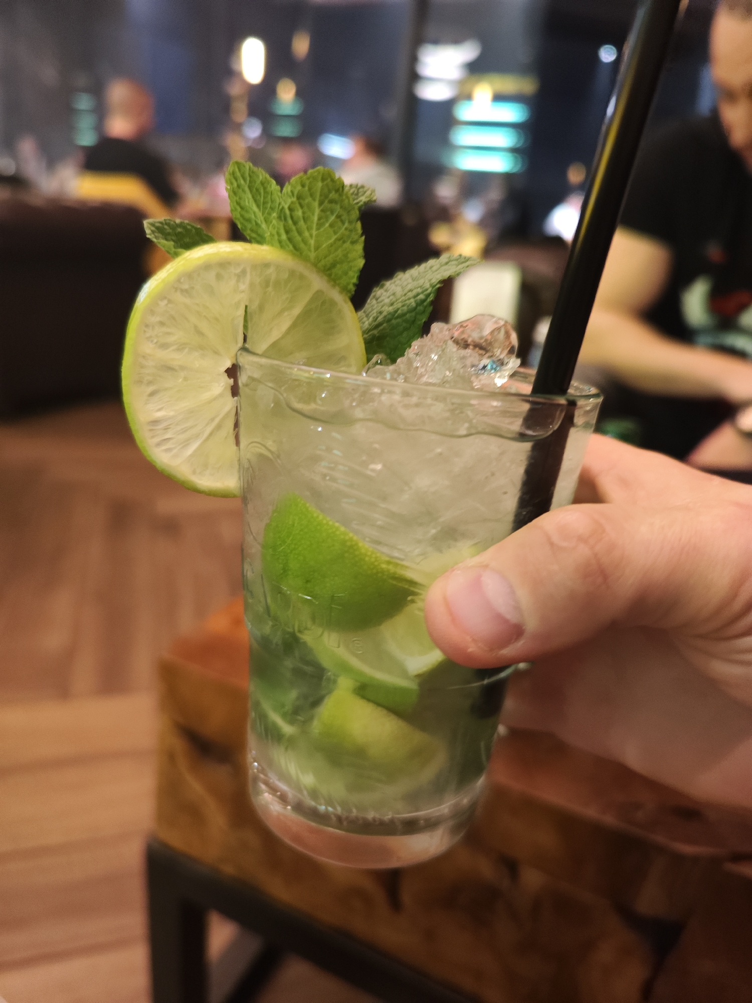 A partially visible adult hand holds a glass of mojito, containing ice, lime slices, and mint leaves, garnished with a lime wedge and mint sprigs. A black straw is inserted into the drink. In the background, blurred figures are seated at tables in a dimly lit indoor setting. The location is potentially in a bar or restaurant in the city of Burgas, Bulgaria. The drink is positioned against a backdrop of wooden furniture and ambient lighting.