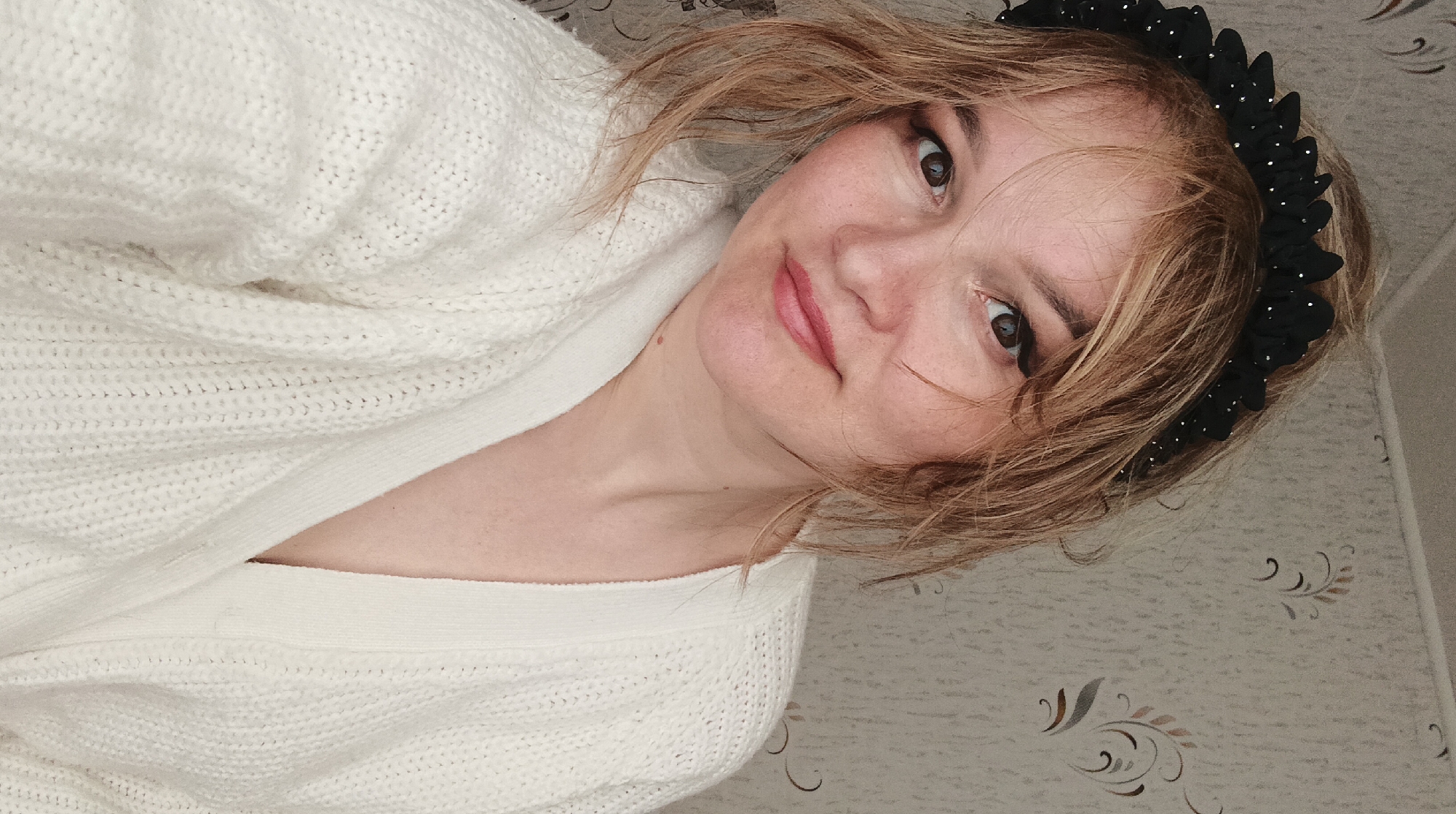 A close-up, chest-up selfie taken indoors in the city of Ольховка, Russia, depicts a light-skinned adult female. She has light brown, slightly disheveled hair, parted to her left, with strands across her forehead. Her brown eyes are visible with dark eyeliner, and her lips are tinted pink, showing a subtle smile. She wears a black fabric headband adorned with small, reflective silver dots, and a white, textured knit cardigan with a V-neck. The background is a wall covered in light-colored wallpaper featuring a repeating pattern of dark, stylized scroll or floral motifs. The lighting is soft and even.