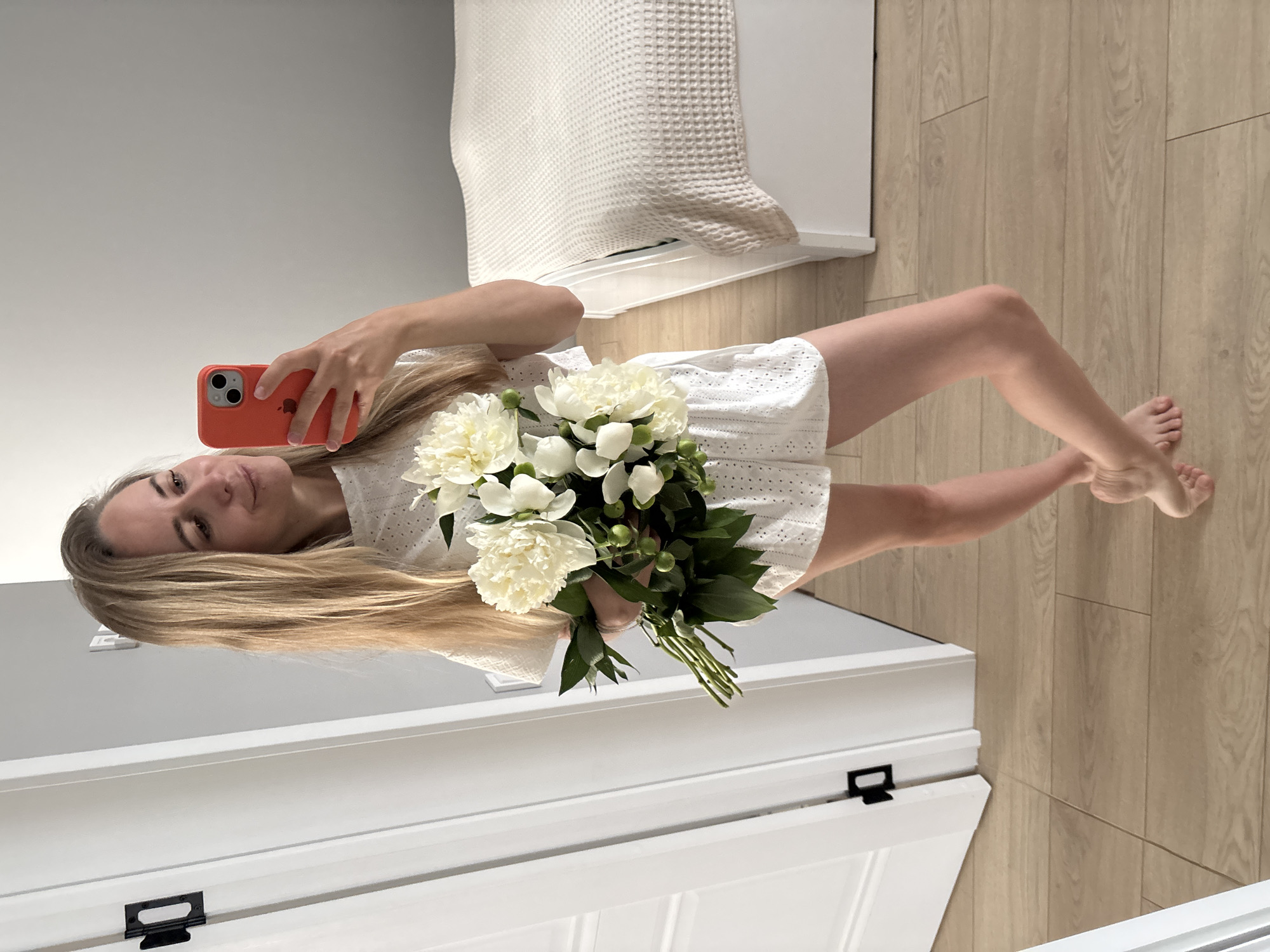 A woman with long blonde hair is taking a mirror selfie while holding a bouquet of white peonies. She is wearing a white eyelet top and shorts. The background appears to be a bedroom with a bed and a white door. The floor is made of light-colored wood. The woman is barefoot. The image was likely taken in a residential setting, potentially in Chaikovsky, Russia, as indicated by the context.