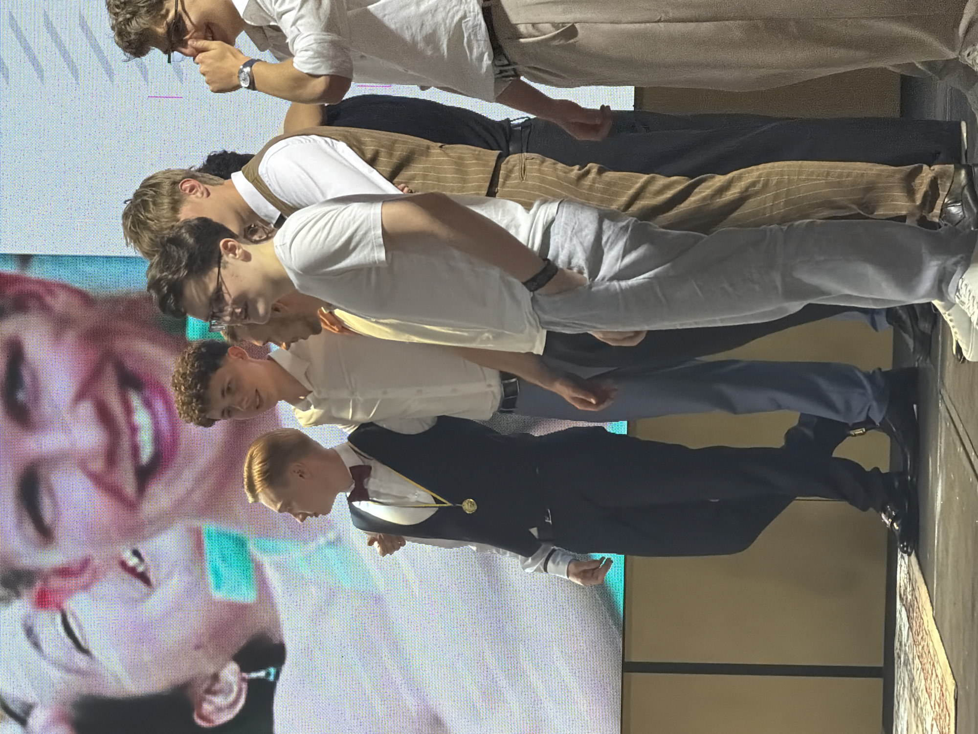 The image shows a group of young men in formal attire standing in a line. They are all facing the same direction, with their bodies angled slightly forward as if presenting or being judged. The man at the front of the line is wearing a dark suit with a white shirt and a burgundy bow tie, and a gold medal is draped around his neck. The man behind him is wearing a white shirt and light-colored pants, and the man behind him is wearing a brown pinstripe vest over a white shirt and dark pants. The man at the back of the line is wearing a white shirt and beige pants. The background appears to be a stage with a large screen displaying an abstract image of a face. The lighting is bright, suggesting an indoor event. The expressions on the faces of the men are varied, but most seem to be focused and determined. There is no visible text in the image.