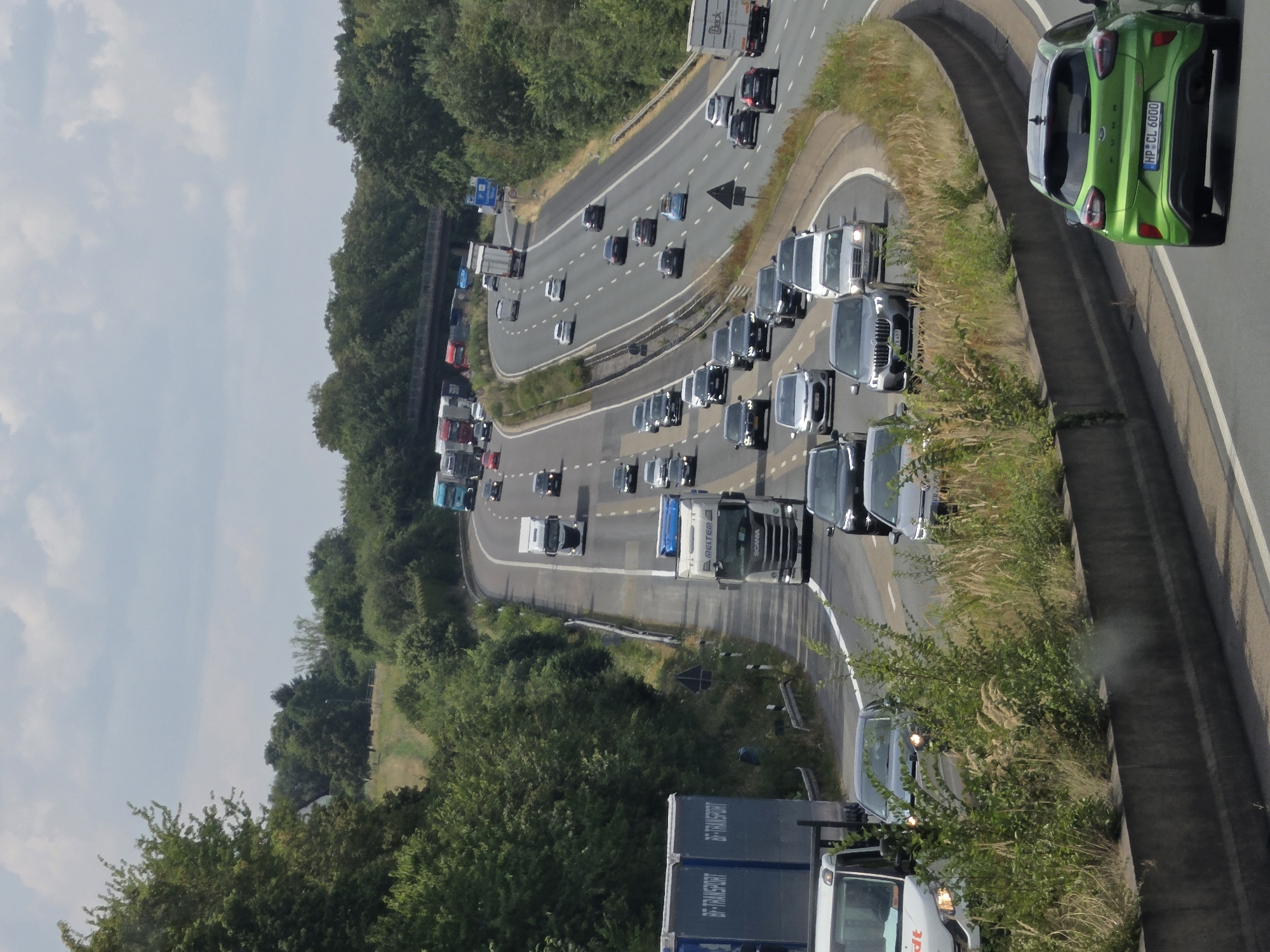 The image captures a view from an elevated position overlooking a multi-lane highway, likely the Autobahn A3, near Bad Honnef, Germany. The highway is congested with traffic, featuring numerous cars and trucks traveling in both directions. On the right side of the highway, a green Ford Puma is prominently visible in the foreground, displaying a license plate that reads "HP CL 6000". The highway curves to the right, bordered by lush green trees and vegetation on both sides. In the distance, more vehicles and a bridge crossing can be seen. The sky is partly cloudy, suggesting an overcast day.