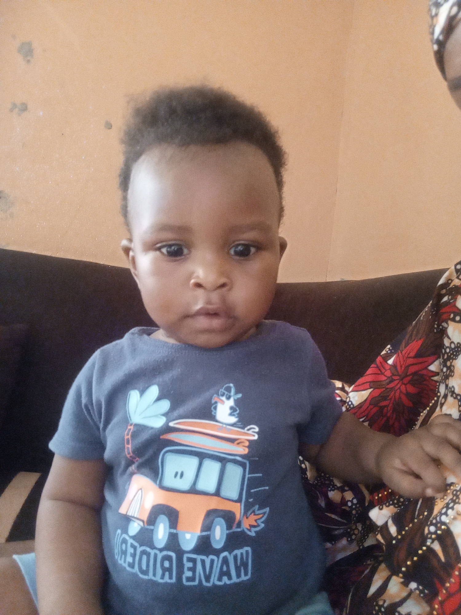A baby wearing a blue t-shirt with a bus graphic is sitting on a dark brown couch. A person with a patterned headscarf and clothing is sitting next to the baby. The background is a plain wall of light orange. The city and location are unknown.