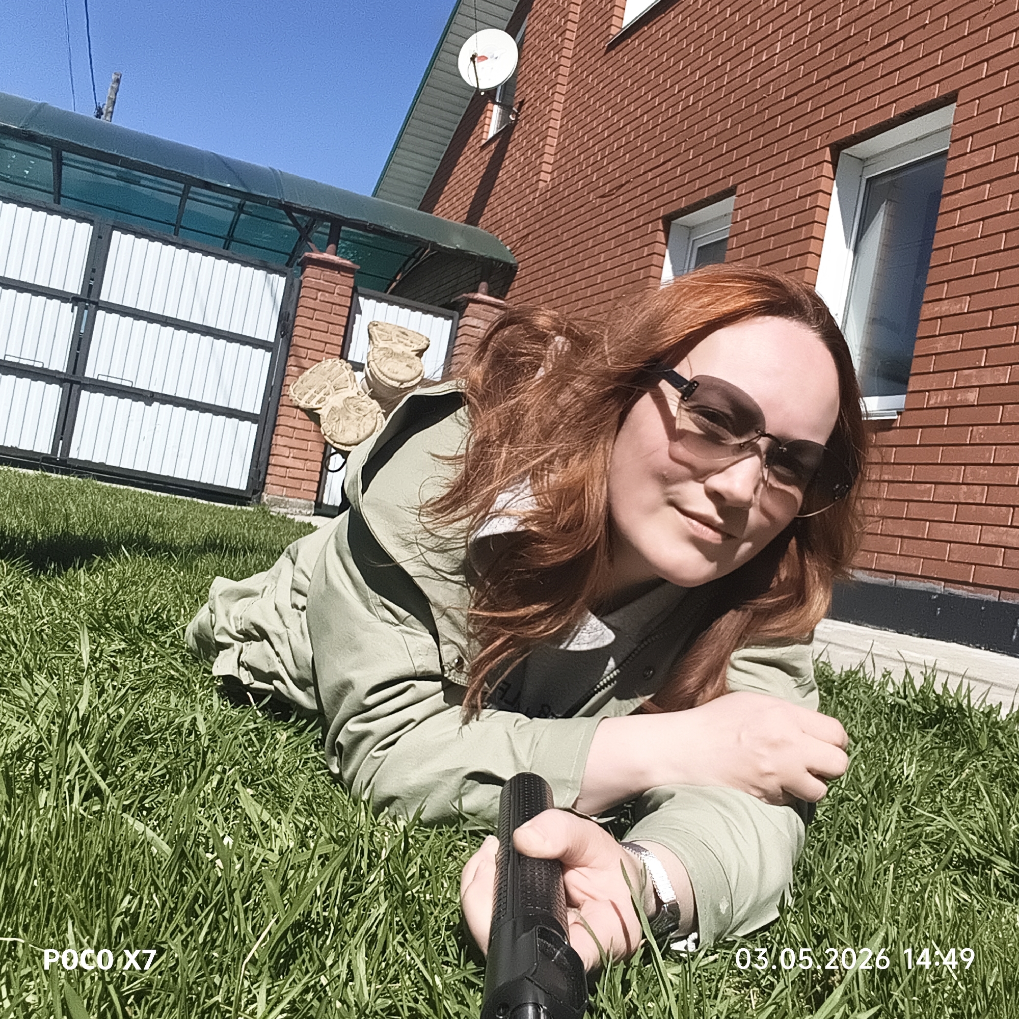 The image depicts a red-haired woman lying on a grassy lawn, holding a black selfie stick. She is wearing sunglasses and a light green jacket. Behind her is a red brick building with white-framed windows and a satellite dish. To the left is a white metal gate with a green awning. Located to the left of the gate is what appears to be concrete lawn decor resembling stacked shoes. The image includes a timestamp of "03.05.2026 14:49" and the camera model "POCO X7." Based on the coordinates, the location is in the city of Chaikovsky, Russia.