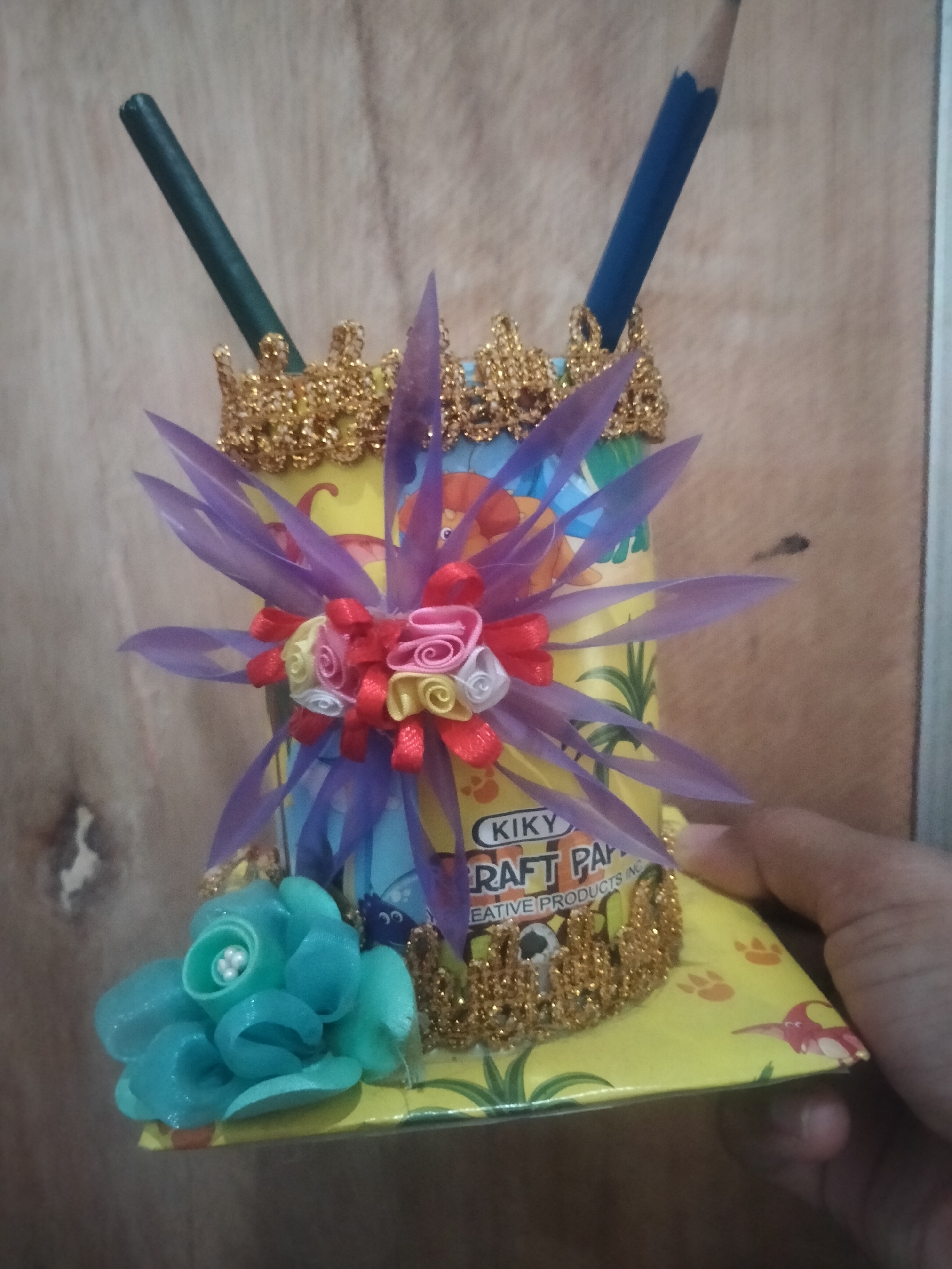 A handmade pencil holder crafted from a yellow paper tube, decorated with golden glittery trim around its top and base, is the central subject. It holds two pencils: one dark green on the left and a blue one on the right. A large, translucent purple flower-like adornment sprouts from the top of the tube, featuring smaller rosettes of pink, yellow, and white ribbon at its center. To the left of the tube, a teal fabric flower with pearl-like beads in its center is attached to the holder's base. The holder itself sits on a yellow, flat surface adorned with paw prints and a cartoonish red bird. A human hand is visible holding the lower right edge of the base. The background is a light brown, textured surface, possibly a wooden wall. The image was taken in Suko, Indonesia.