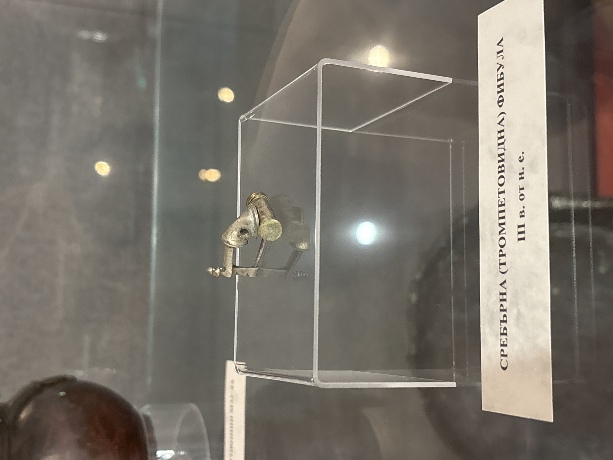 The image displays a silver, trumpet-shaped fibula, an ancient brooch, presented in a museum setting. The artifact is small, made of tarnished, silver-colored metal, and rests on a clear acrylic stand inside a clear acrylic display case. Reflections from the surrounding environment, including blurred light sources, are visible on the case's surfaces.
To the right of the display, a white informational sign stands vertically. The text, written in Cyrillic, identifies the object. It reads: "СРЕБЪРНА (ТРОМПЕТОВИДНА) ФИБУЛА," translating to "Silver (trumpet-shaped) fibula." Below this, "III в. от н. е." indicates the artifact dates to the "3rd century AD."
The background is dark and out of focus, featuring soft, blurred circles of light, characteristic of an indoor museum exhibit with selective lighting. No people are visible, and the scene is static, showcasing the historical artifact. The context suggests the location is likely a museum in Devnya, Bulgaria.