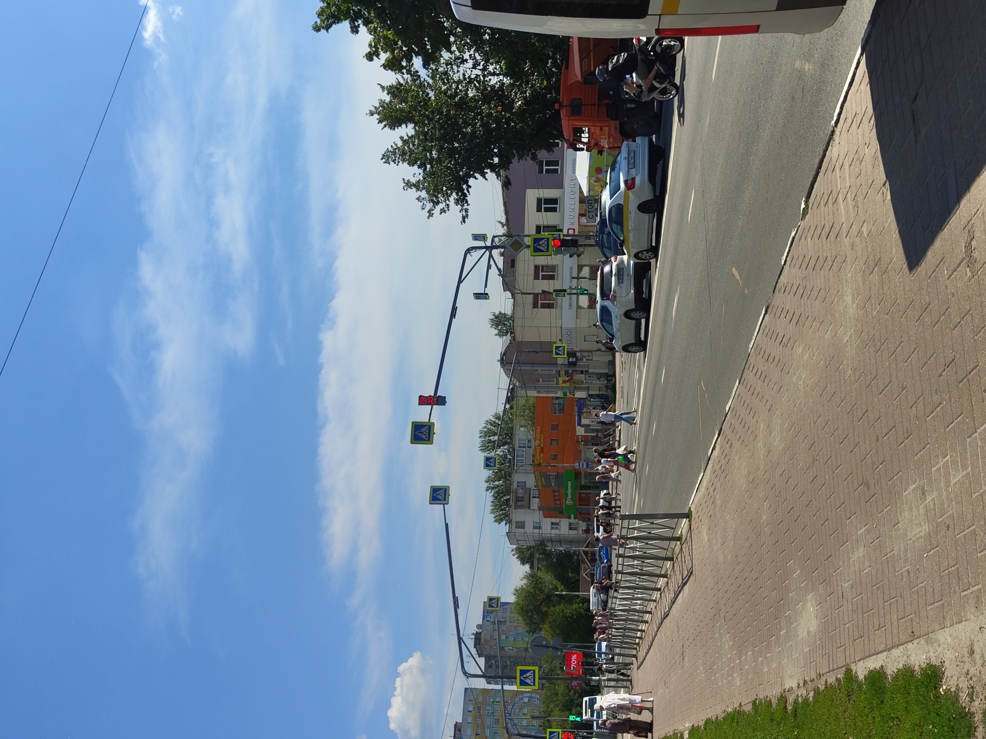 A wide, paved pedestrian walkway runs along the left side of a multi-lane asphalt road in Ramenskoye, Russia, under a bright blue sky with scattered white clouds. A metal fence separates the walkway from the road. A grassy strip borders the far left of the walkway. On the walkway, two elderly women, one in a white outfit and the other in a brown outfit, are walking from left to right.
The road is busy with vehicular traffic. Several cars are stopped at a traffic light, including a white SUV and a white sedan with a yellow stripe. A large orange truck is also present, and a person on a motorcycle is riding past it. Pedestrians are crossing the road at a marked crosswalk. Various buildings are visible on the right side of the road, including multi-story apartment buildings and smaller commercial establishments. Traffic signals and pedestrian crossing signs are mounted on poles above the road. The scene appears to be a typical urban intersection on a sunny day.