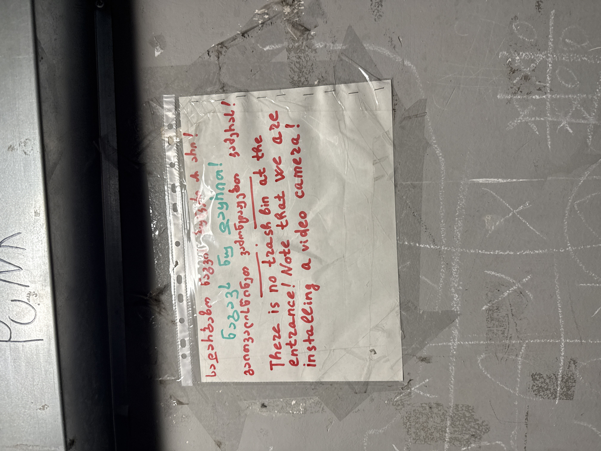 A handwritten note is affixed to a wall, covered in a clear plastic sleeve. The note is written in red ink with some lines in teal. It appears to be in two languages, Georgian and English. The Georgian text reads: "სადარბაზოს ნაგავი გვაქვს, ნაგავს ნუ დავყრით, გათავისუფლებით ვამთავრებთ, გამოყენებამ". The English text reads: "There is no trash bin at the entrance! Note that we are installing a video camera!". The wall is a rough, grey concrete surface with chalk markings, including what appears to be a tic-tac-toe grid. Above the note, on a metal surface, the word "PUNK" is written in black marker. The overall environment suggests an outdoor or semi-outdoor public space, potentially a stairwell or alleyway, in Tbilisi, Georgia. The lighting is dim.