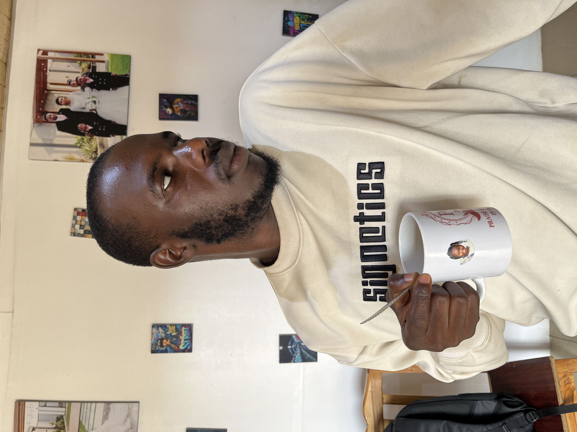 The image shows a man sitting in a room. He is wearing a light-colored sweatshirt with black text on it. He is holding a white mug in his right hand and what appears to be a spoon in his left hand. There are several framed pictures on the wall behind him, including what looks like a wedding photo and some smaller, colorful artwork. The lighting suggests it might be daytime. The man appears to be looking upwards and to the left. The word "SIGNETICS" is visible on his sweatshirt. The mug has some text and images on it, including a smaller circular picture of a child and the words "PRAISE". The room appears to be indoors, possibly a home or office, with wooden furniture visible.