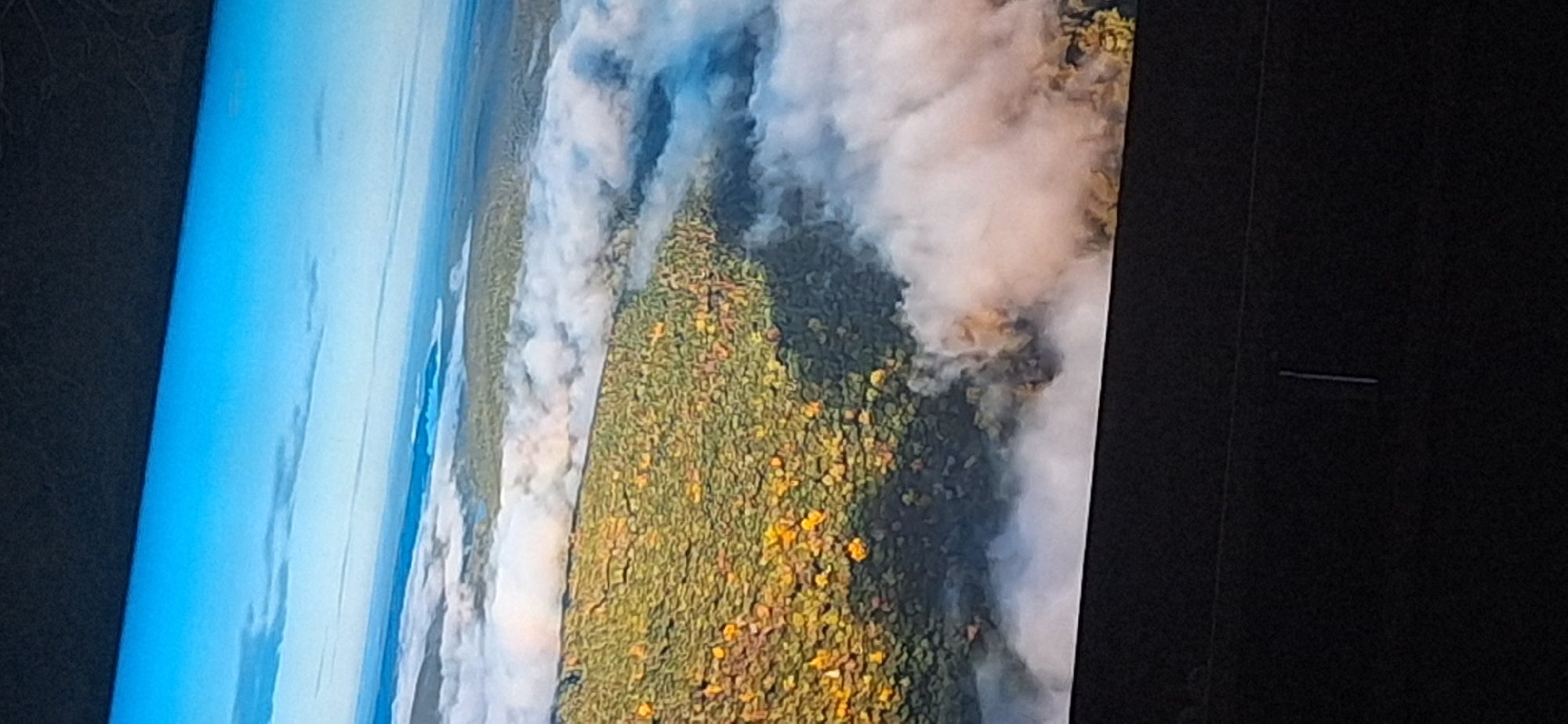 The image displays an aerial view of a forest landscape partially obscured by fog or clouds. The trees exhibit autumn foliage with patches of yellow and green. The sky is clear blue with some wispy clouds. The foreground shows a dark, out-of-focus area, likely the edge of a screen or frame, and a faint vertical line. There are no discernible people, animals, buildings, or specific landmarks that would indicate a location in Bila Tserkva, Ukraine.