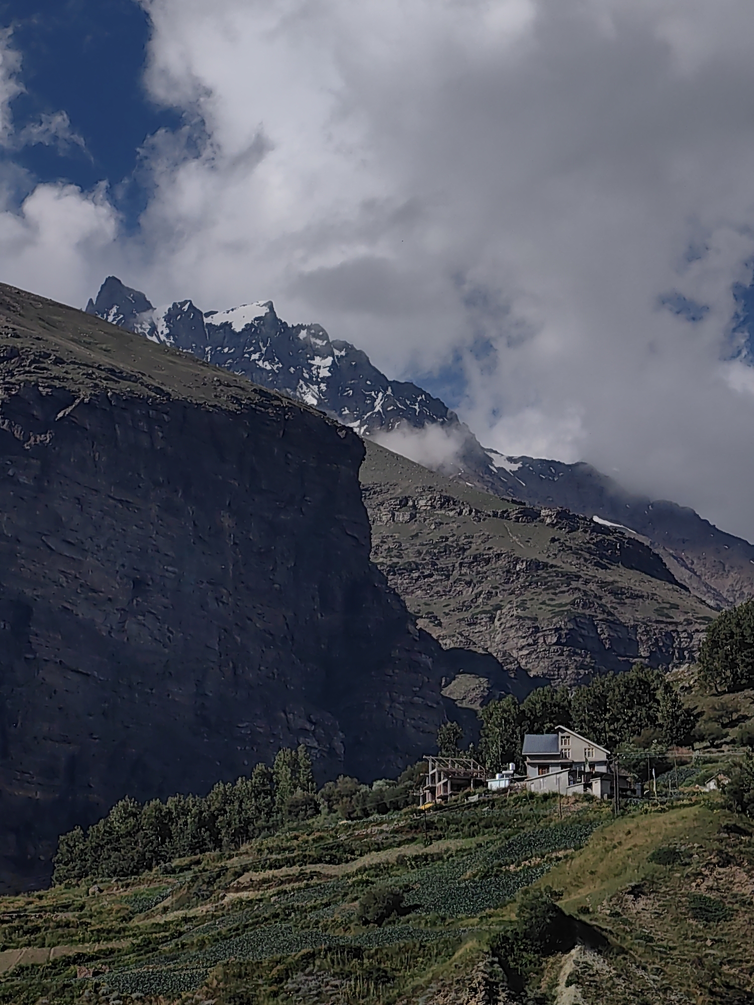 The image depicts a mountainous landscape with a village nestled on a terraced hillside. In the foreground, lush green vegetation covers the slopes, with visible patches of crops. Several buildings, appearing to be houses and possibly a construction site, are situated on the hillside, some with visible roofing and windows. Tall, dark green trees line the upper portion of the village area. The midground is characterized by rugged, rocky mountain slopes with patches of greenery and some scattered trees. In the background, imposing snow-capped mountains rise majestically, with dramatic clouds obscuring parts of their peaks. The sky features a mix of blue patches and white clouds. The overall scene suggests a remote, high-altitude location, likely in the Himalayas, given the mention of Yamling, India. The image is captured in daylight, with shadows indicating the direction of the sun. There are no visible people or animals in the frame.