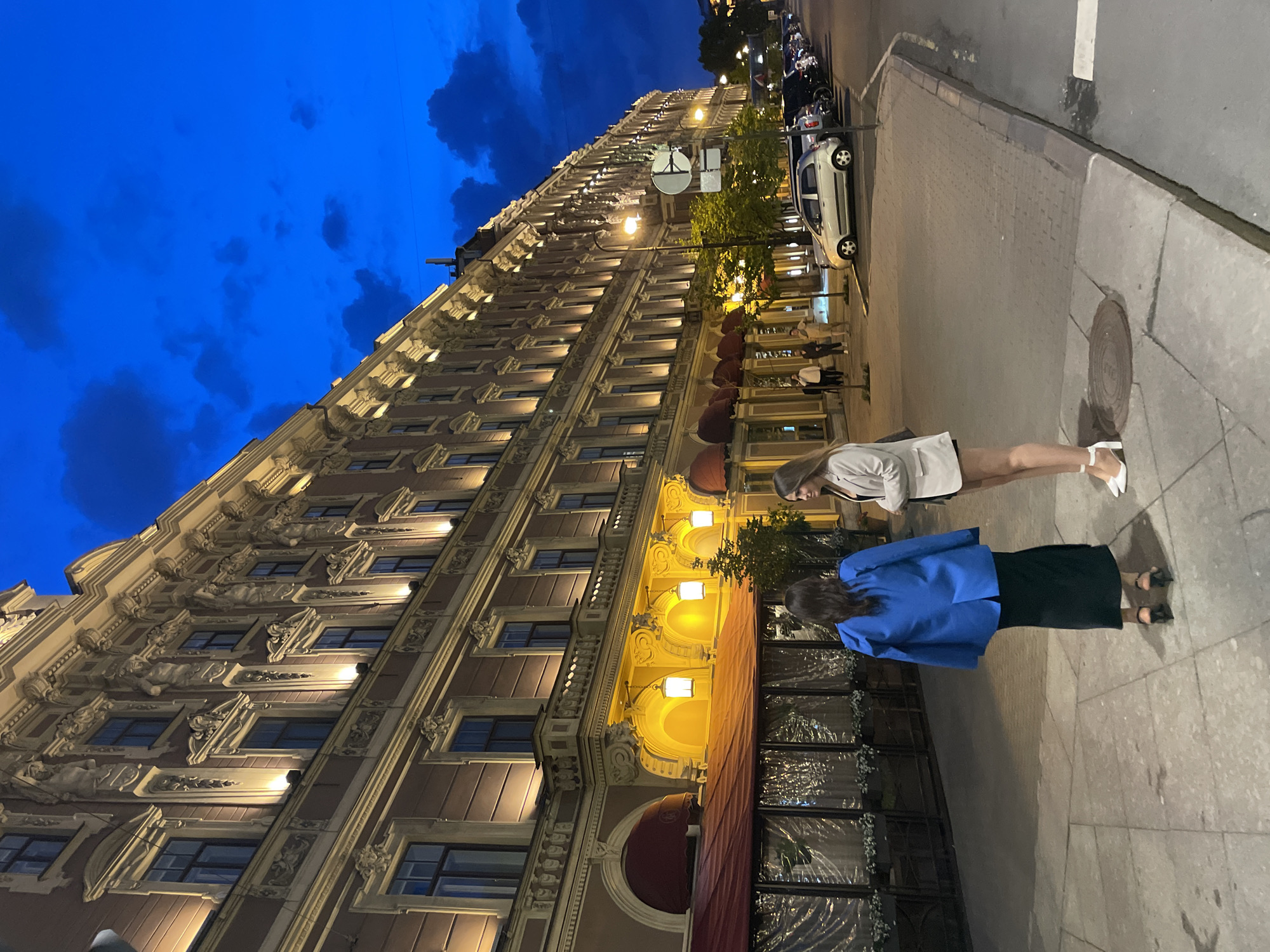 The image captures an evening or night scene on a city street in Saint Petersburg, Russia, dominated by a long, multi-story historic building. The building facade, light brown with cream-colored architectural detailing, features numerous windows with ornate surrounds, pilasters, balustrades, and statuary elements, all illuminated by warm upward-facing lights. The ground level exhibits a series of arched entries and storefronts, some with red awnings. One section features an enclosed outdoor dining area with transparent plastic sheeting. Ornate pendant lanterns hang from the archways, casting a warm glow.
The sky above is a deep blue with scattered dark clouds, indicative of twilight or early night.
On the paved stone sidewalk in the foreground, two women are standing. The woman on the left, facing away from the camera, has dark hair and wears a royal blue oversized blazer over a long dark skirt and dark high-heeled shoes. The woman on the right, in profile facing the first woman, has long light brown hair and wears a light beige blazer, dark shorts, and white high-heeled shoes with ankle straps. She holds a small object in her hands.
Further down the street, additional indistinct figures are visible walking on the sidewalk. A row of parked vehicles, including a light silver compact car, occupies the street parallel to the curb on the right side. Several small trees with green foliage are planted along the sidewalk, and a circular white sign on a pole is visible on the right. A circular manhole cover is embedded in the sidewalk near the center foreground.