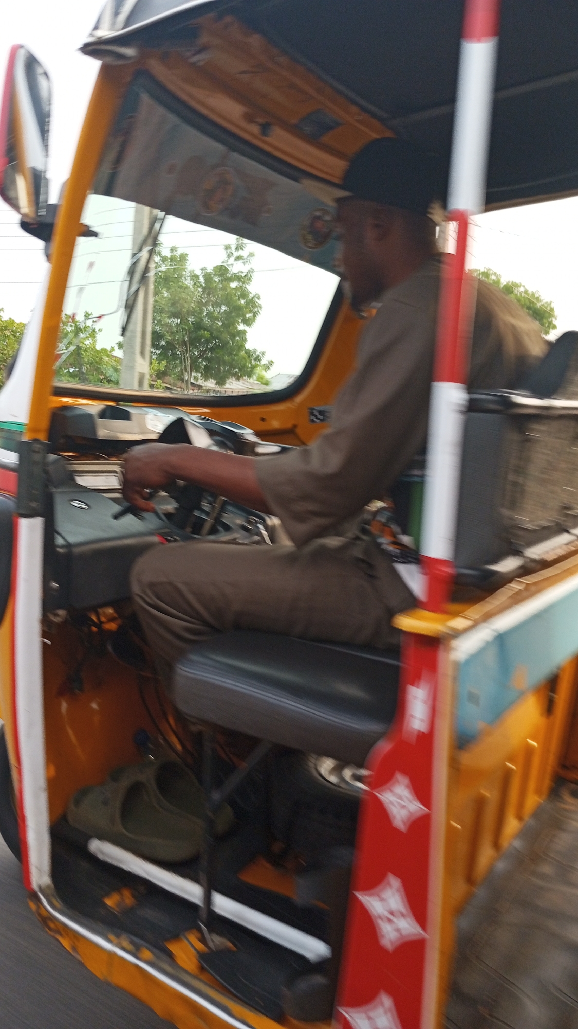The image shows the interior of a yellow tricycle taxi, with a Black male driver wearing a brown shirt and black cap operating the vehicle. The taxi has a red and white patterned bar near the passenger area. On the floor of the taxi, a pair of green sandals are visible. The background shows a street scene with trees and buildings, suggesting an urban environment. The location is Zango-ndaji, Nigeria.