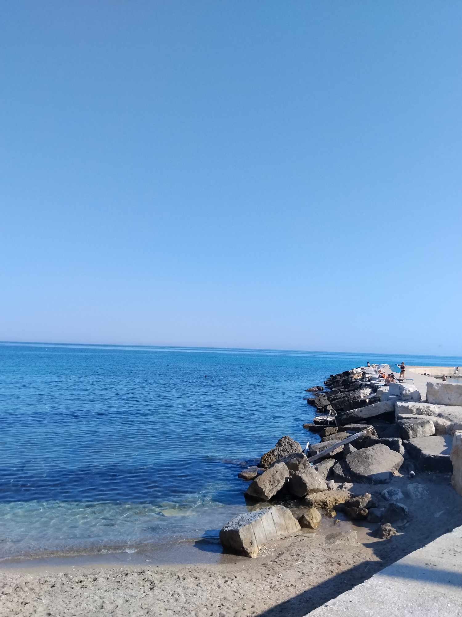 The image shows a serene coastal scene. The sky is a clear, uninterrupted blue, meeting the horizon of a calm, deep blue sea. The water is transparent near the shore, revealing the sandy bottom. A man-made structure composed of large, rough-hewn stones and concrete blocks juts out into the sea, creating a breakwater or pier. Several people are visible on this structure, some standing and others seated, appearing to be enjoying the day. One person on the pier is holding a long object, possibly a fishing rod or a camera. The location is identified as the Lungomare Nazario Sauro in Bari, Italy.
