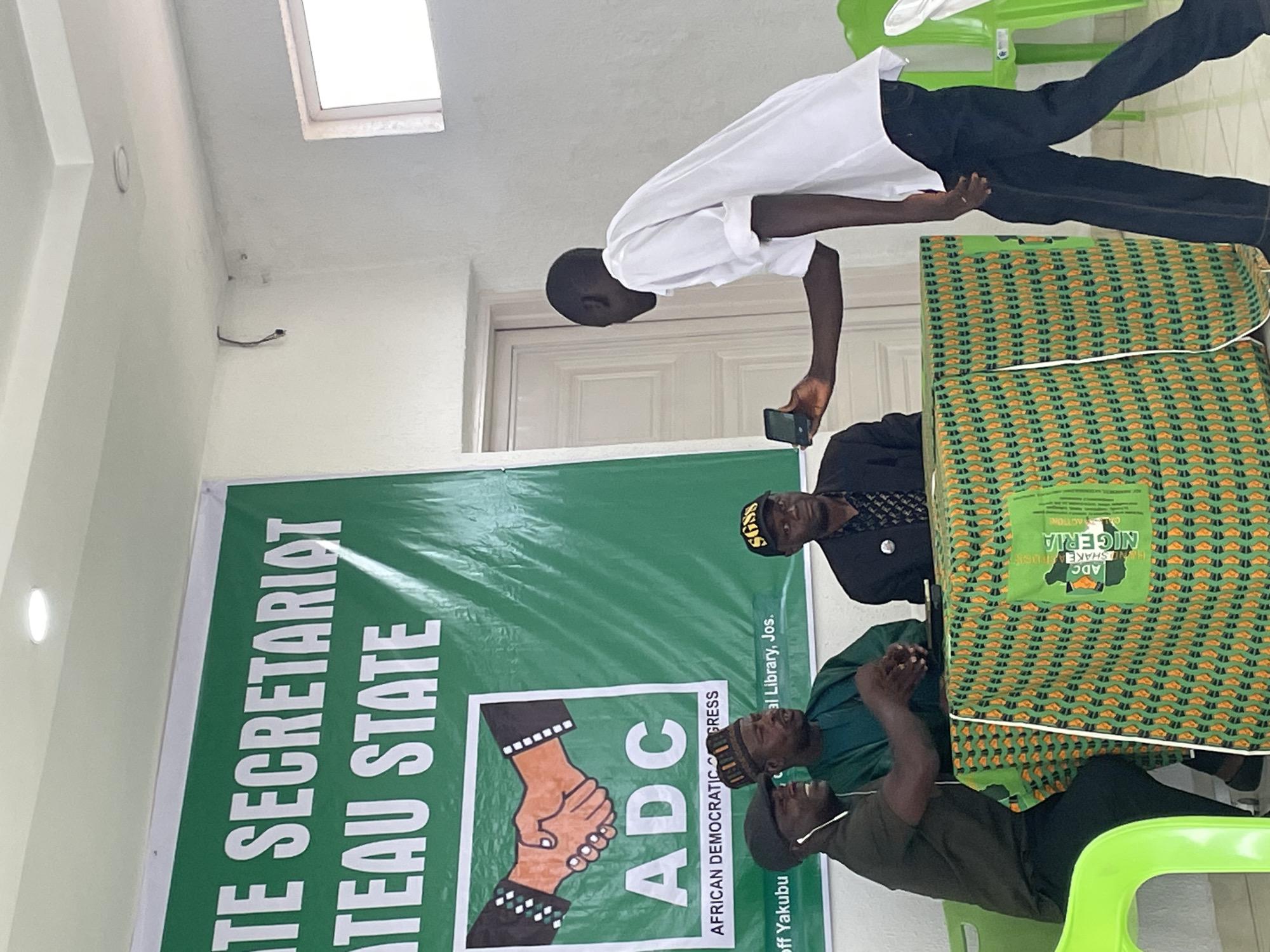 The image shows a gathering of people inside what appears to be a political office or meeting room, as indicated by a banner displaying the acronym "ADC," which stands for African Democratic Congress, and mentions "Plateau State Secretariat." The location is specified as "al Library, Jos," suggesting it's situated in or near a library in Jos, Nigeria.
Three men are seated behind a table draped with a patterned cloth. One man is standing beside the table, holding a phone as if taking a picture of the group. Another man wears a black hat with the letters SGSS on it, indicating a possible association or affiliation. Other men seated behind the table are wearing traditional Nigerian attire. The room is sparsely furnished with light-colored walls and green plastic chairs. A banner with the "ADC" logo and the slogan "UNDISHAKE SHAKEARRIN A RUISE ACTIONE" is visible in the background. The lighting is bright, possibly natural light from a window.