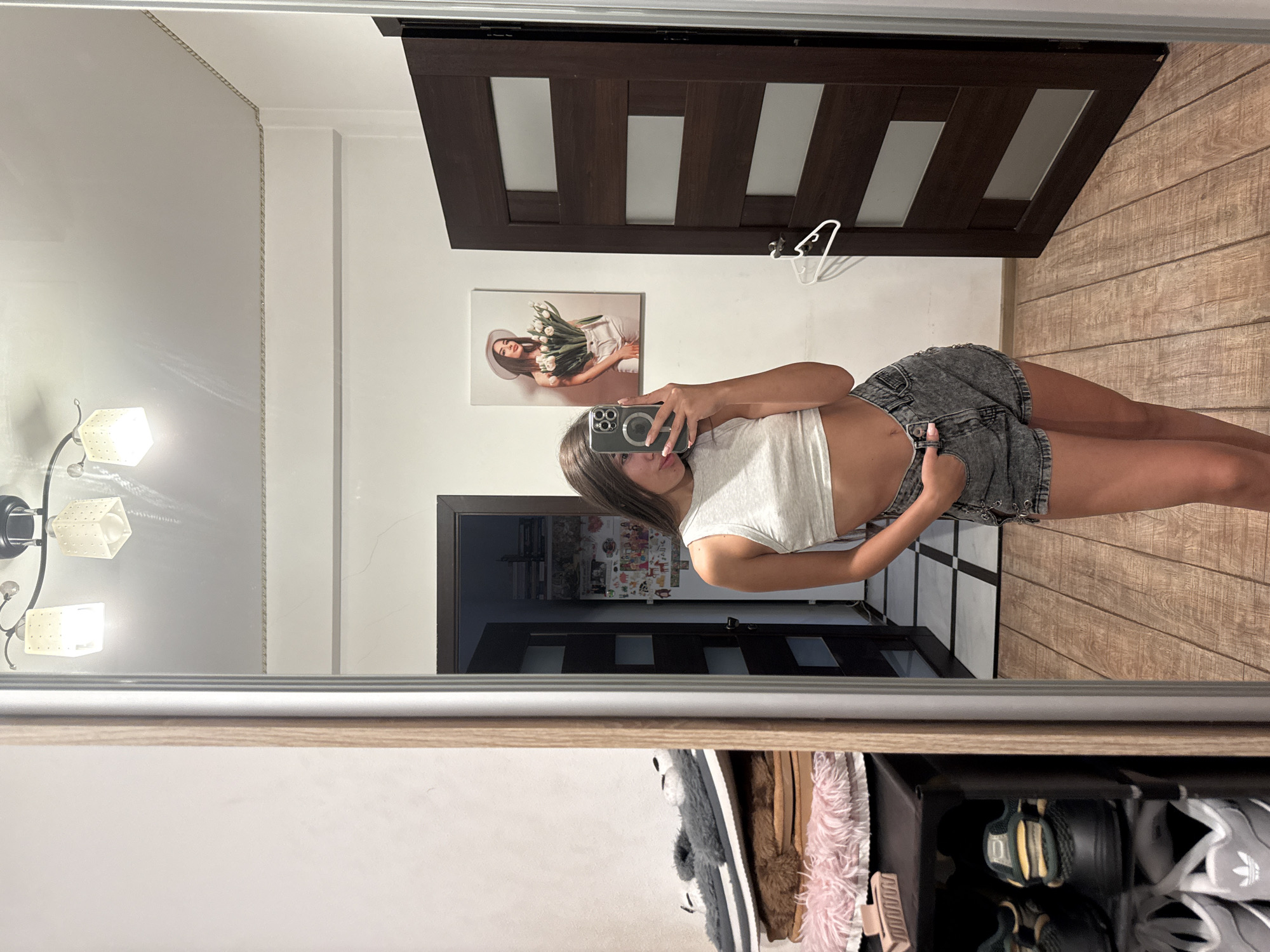 A young woman with long brown hair is standing in a room, taking a selfie in a large mirror. She is wearing a light grey crop top and distressed denim shorts. Her left hand is in her pocket and she is biting her lower lip. In the reflection of the mirror, a dark wooden door is visible, with a white clothes hanger hanging on it. To the left of the door, a framed picture of a woman holding a bouquet of white tulips is hanging on the wall. In the reflection of the mirror to the left of the woman, a closet is visible, with shoes and slippers stacked on shelves. The floor is made of light brown wood planks. The room appears to be an apartment in Chișinău, Moldova.