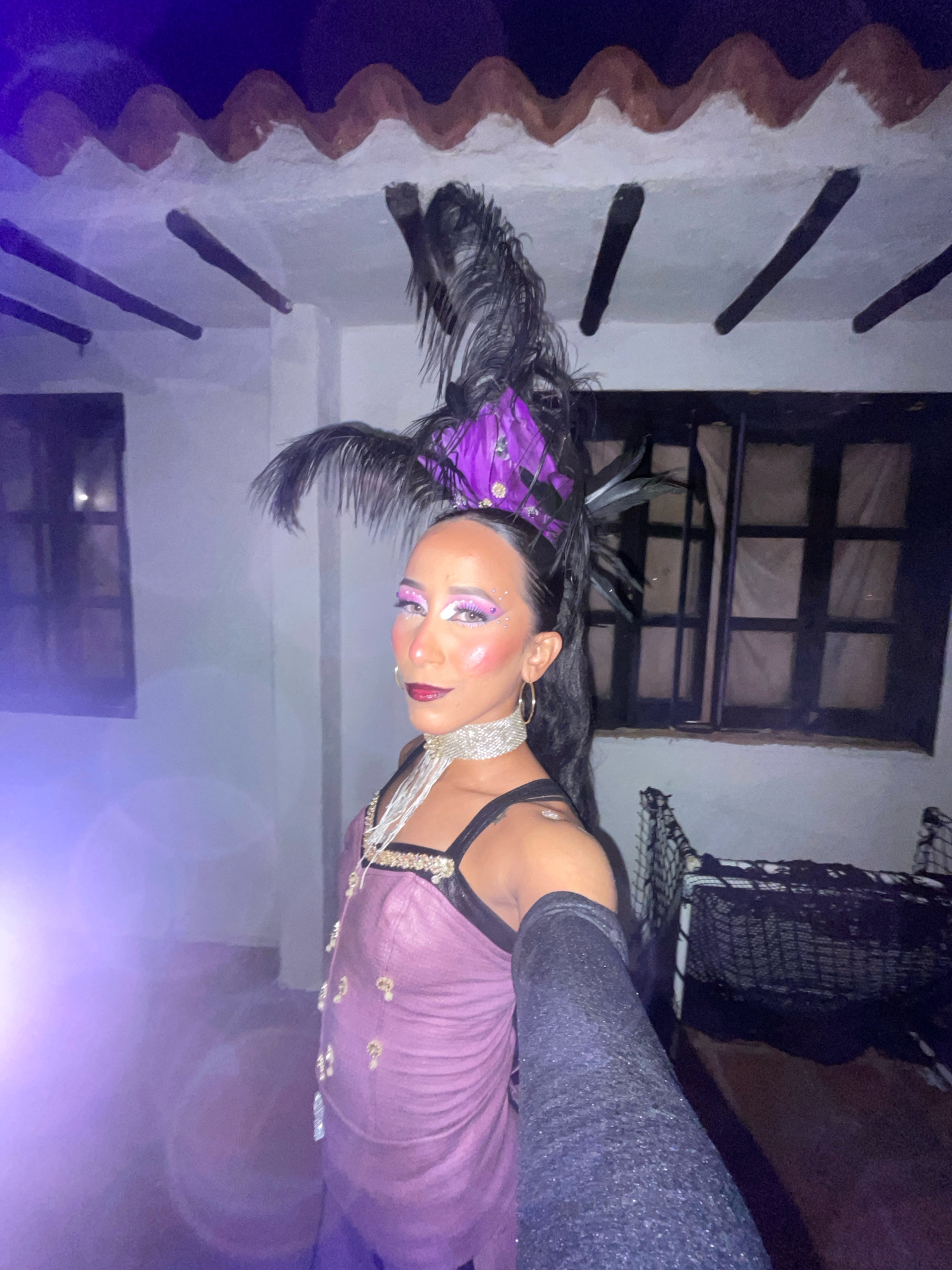 A person in a costume, likely for a performance or event, poses for a selfie outdoors at night. The person wears a theatrical headdress with purple and black feathers, dramatic eye makeup with glitter accents, and a shimmering purple gown adorned with gold embellishments. They also wear a sparkling choker necklace and long, dark sleeves. The background appears to be a courtyard or patio area with a white-washed wall, dark wooden beams supporting a tiled roof, and a dark-paned window. A black net structure is visible to the right. The lighting suggests a festive or performance atmosphere, with a purple hue cast on the left side of the image. The scene is likely located in Porlamar, Venezuela, given the architectural style of the building which is characteristic of some older structures found in coastal towns in Venezuela.