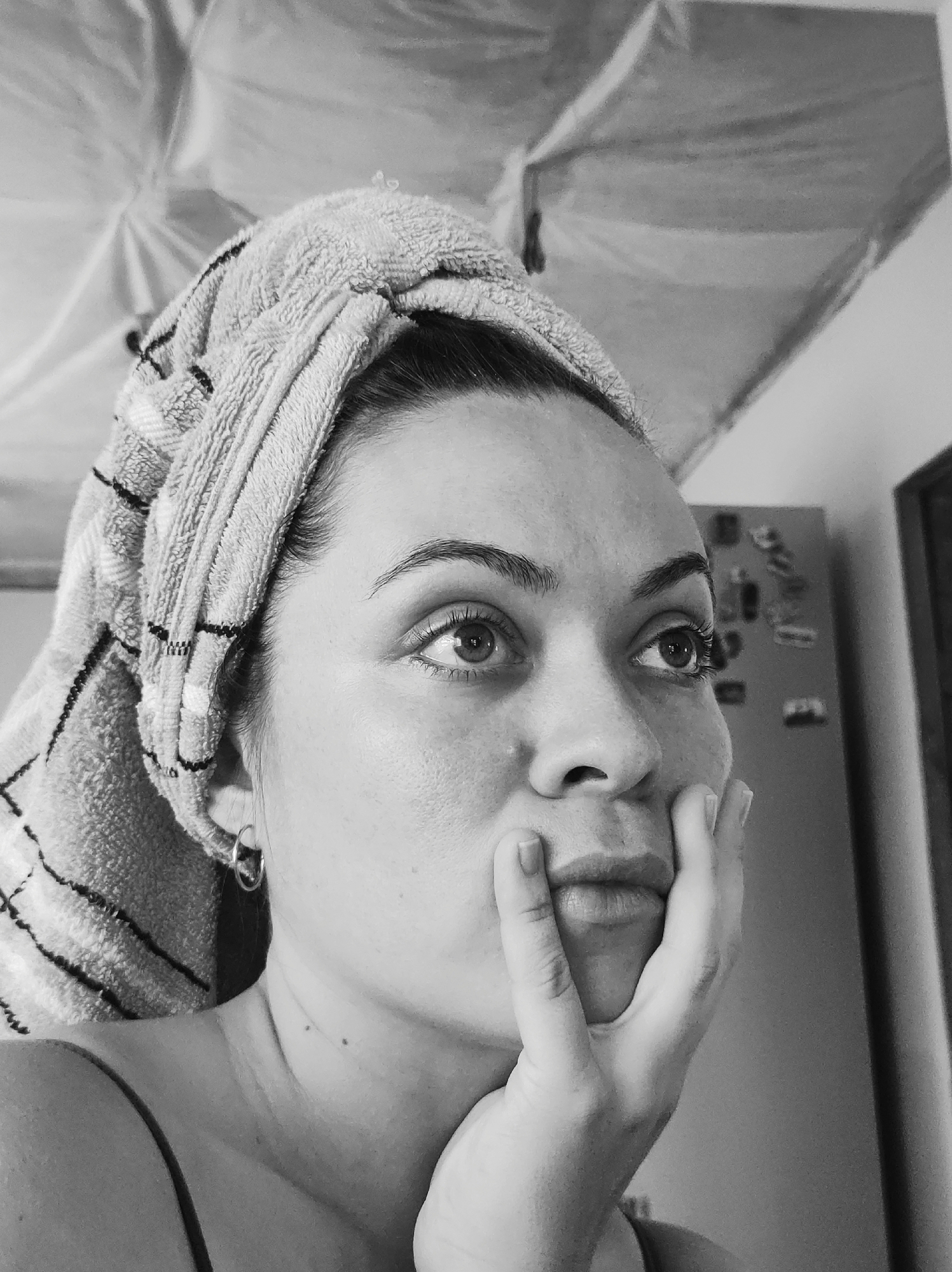 This black and white, close-up portrait features an adult female indoors in Unknown, Unknown. She is positioned in the lower left quadrant of the frame, facing forward but looking upwards and towards her right. A light-colored, striped terry cloth towel is wrapped around her head in a turban style, covering most of her dark hair. Her right hand rests against her chin and lower lip, with fingers slightly splayed. She wears a small, metallic hoop earring in her visible left ear and a thin-strapped garment on her shoulder. Her eyes are open and directed upwards, exhibiting reflections of light. Two small moles are visible on her neck. The background on the upper left consists of a light-toned, draped or folded fabric-like surface. The background on the right reveals part of a dark-colored refrigerator adorned with several small, light-colored magnets, and a darker vertical structure further to the right. The overall lighting is soft and diffuse.