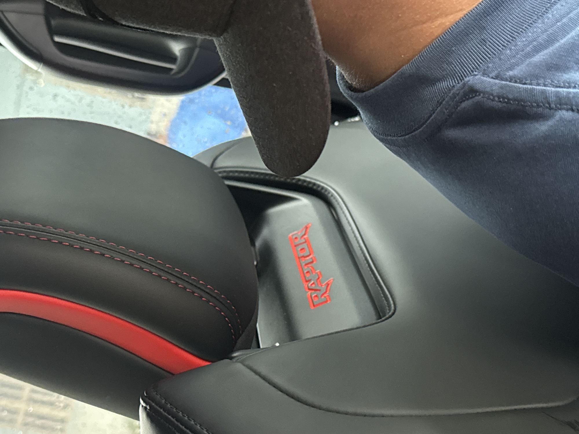 A close-up shot inside a vehicle focuses on the passenger seat, adorned with red stitching and a red accent stripe. The word "RAPTOR" is emblazoned in red on a black surface behind the seat. A gloved hand is visible in the upper left corner, while a person wearing a blue shirt occupies the right side of the frame. The scene is observed from within the car, offering a view of the exterior, seemingly through the windshield. There is no evidence to support the scene's location being Nghia Lo Ward, Vietnam.