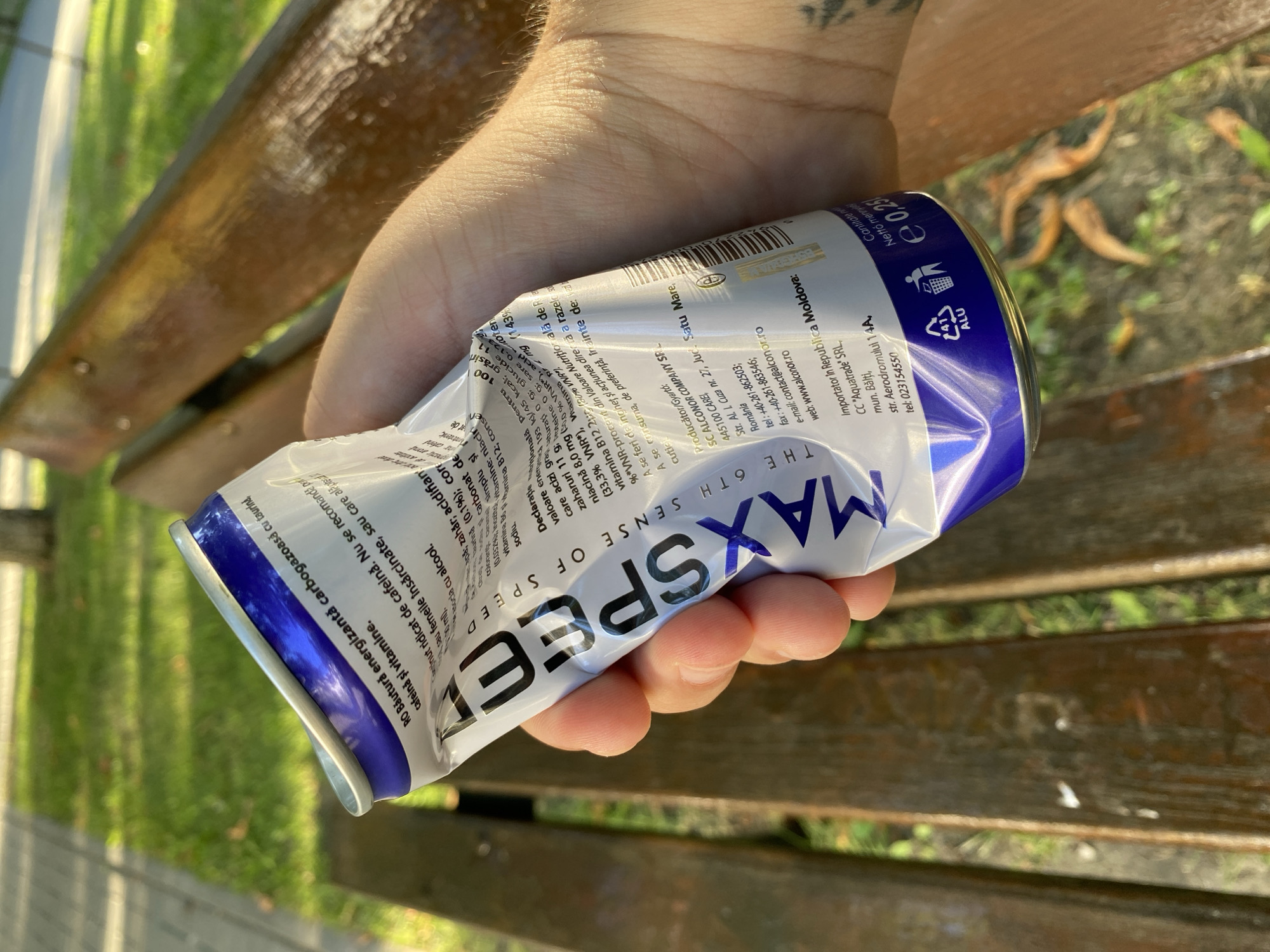 A person's hand holds a crushed aluminum can of a beverage, the label of which appears to be in Romanian. The can has a white and purple design and the text "MAX SPEED" is prominently displayed. The hand is positioned in the foreground, obscuring part of the can. In the background, a wooden park bench is visible, along with green grass and trees, suggesting an outdoor setting. The lighting indicates it is daytime, with shadows cast by the sun. The overall impression is one of a casual outdoor scene, possibly in a park.
This image does not provide any information about the city of Chișinău, Moldova.