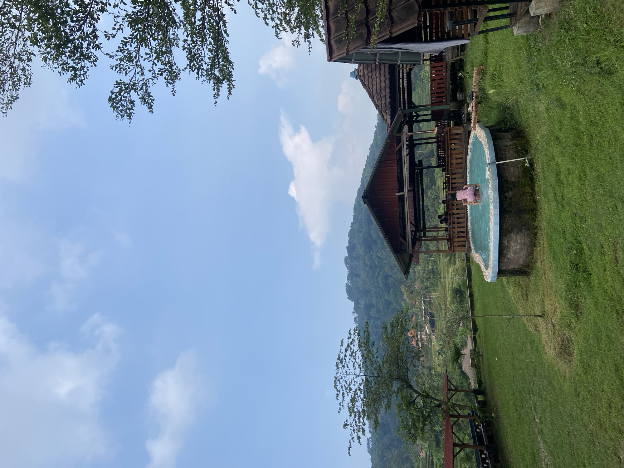 The image shows an outdoor scene near Penanggungan, Indonesia. The view includes a partially obscured mountain in the background and a blue and white tiled swimming pool. A wooden structure with a brown roof is adjacent to the pool. Lush green grass covers the ground. The sky is blue with scattered white clouds. Tree branches frame the top left corner. A person is partially submerged in the pool.