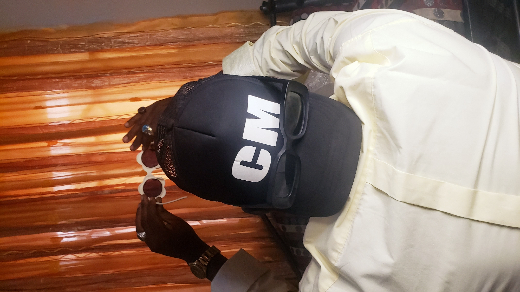 A person wearing a white shirt and a black cap with the letters "CM" in white is holding up a pair of sunglasses with red lenses and white frames in front of a brown and orange striped background. The person is also wearing a watch and rings on their fingers. The location is Narayi, Nigeria.